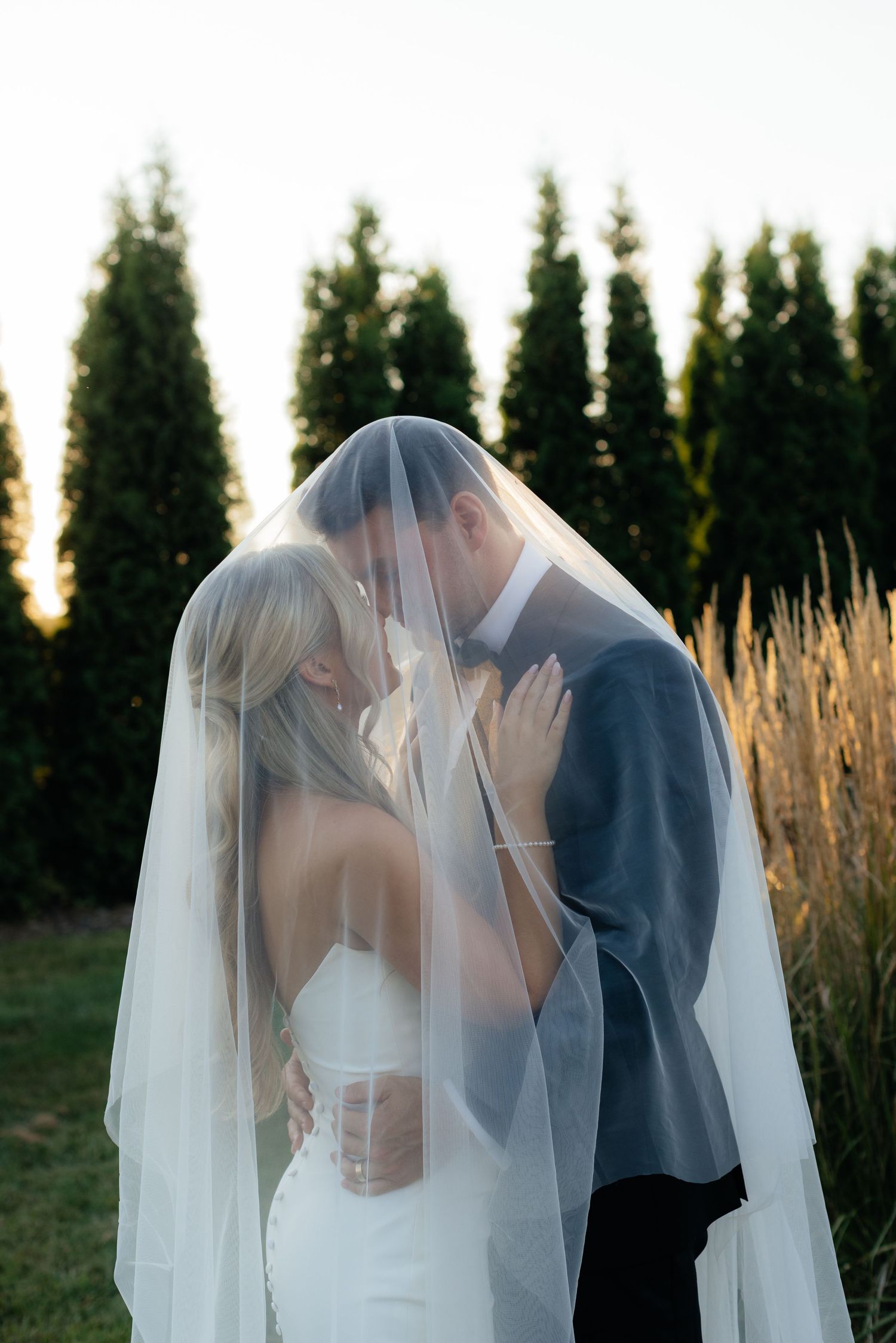 A tender moment between newlyweds under a flowing veil against a sunset backdrop.