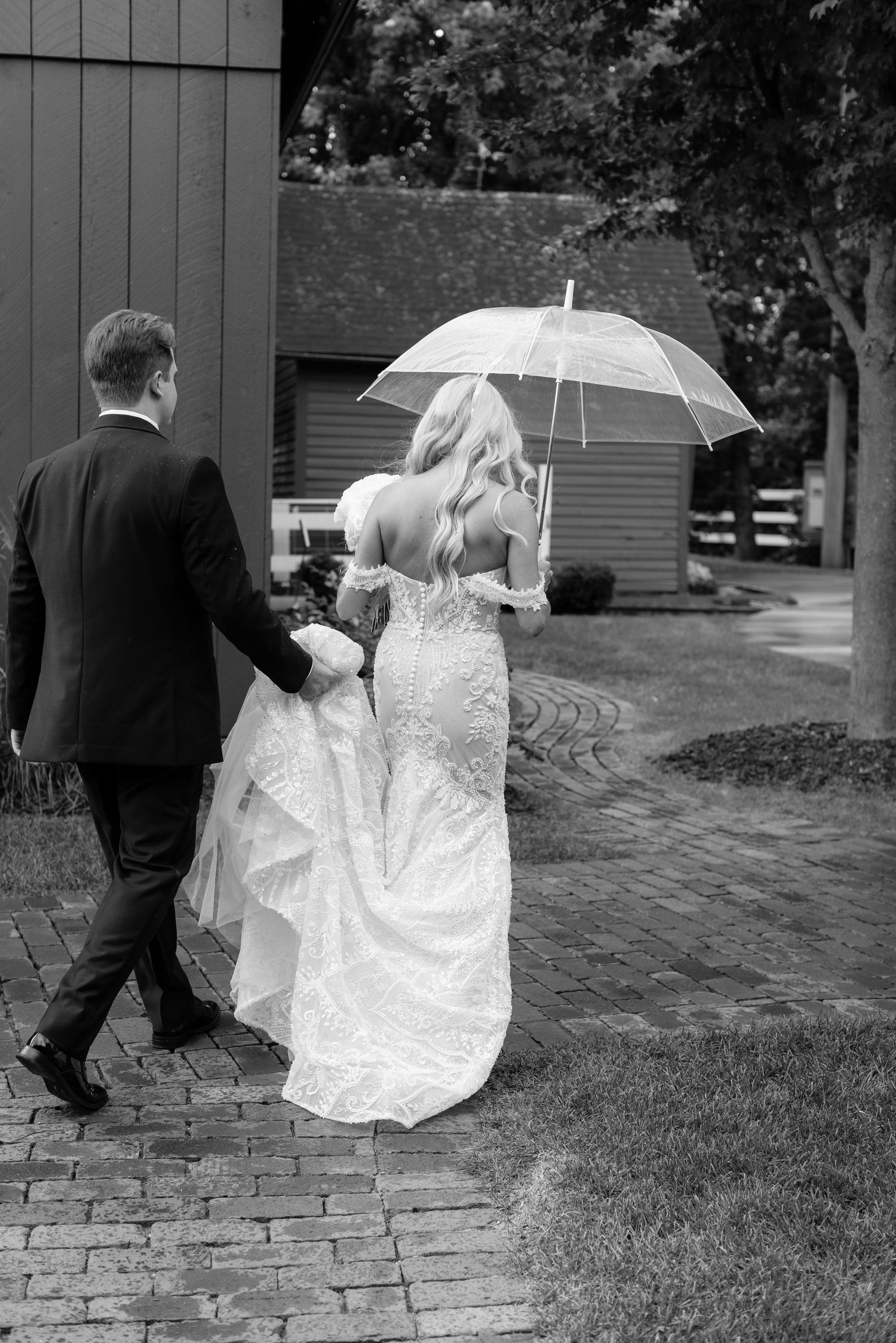Wedding couple walks along brick pathway holding white parasol on overcast day.