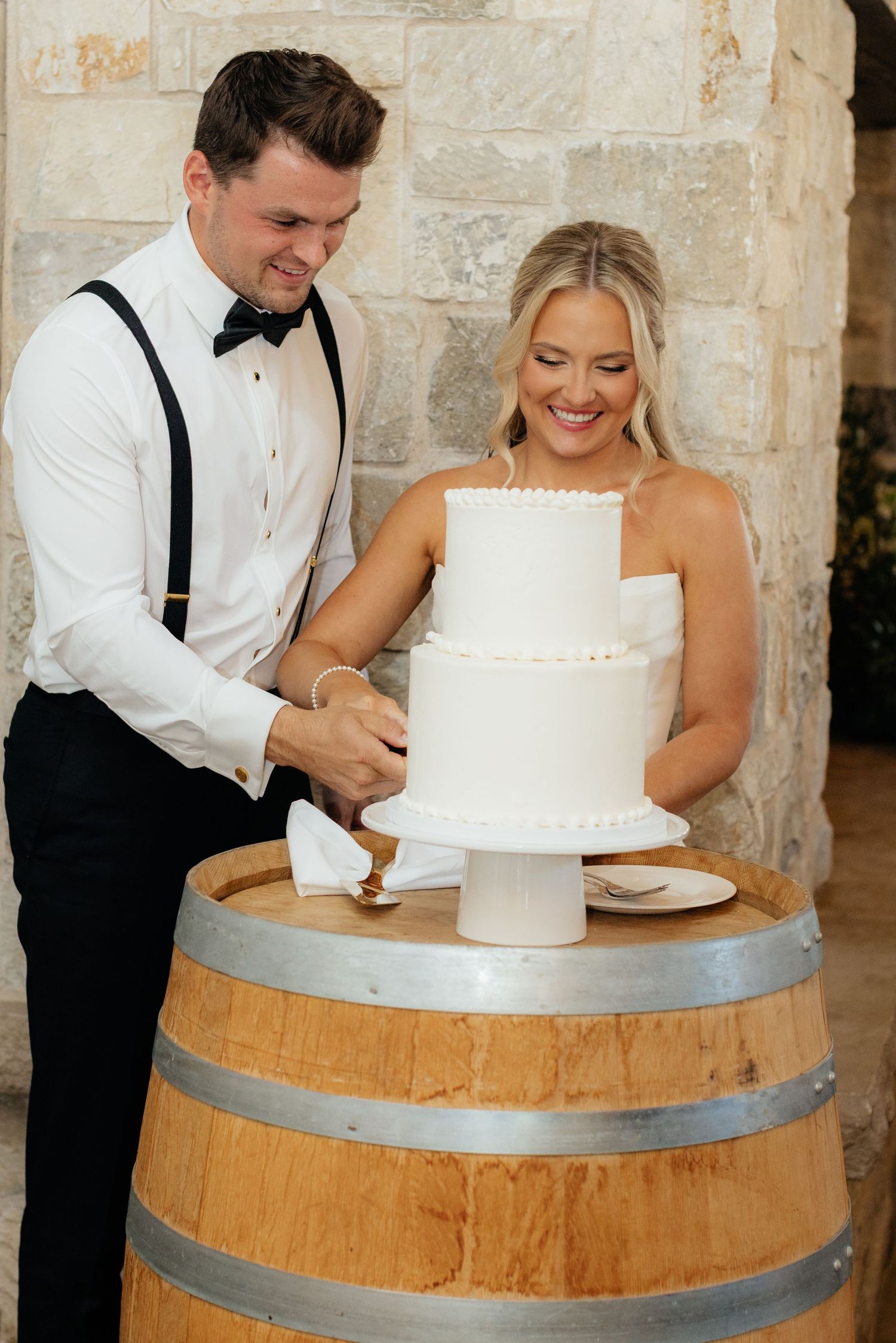 A couple laughing together while preparing to cut their white wedding cake displayed on a rustic wine barrel.