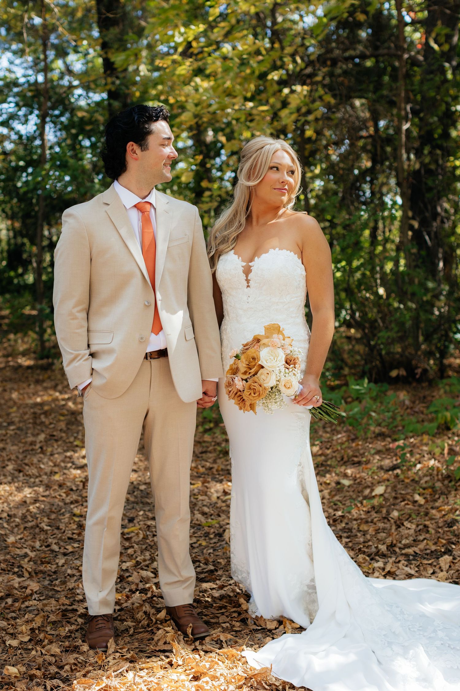 Wedding couple in tan suit and white dress pose together among fallen autumn leaves.