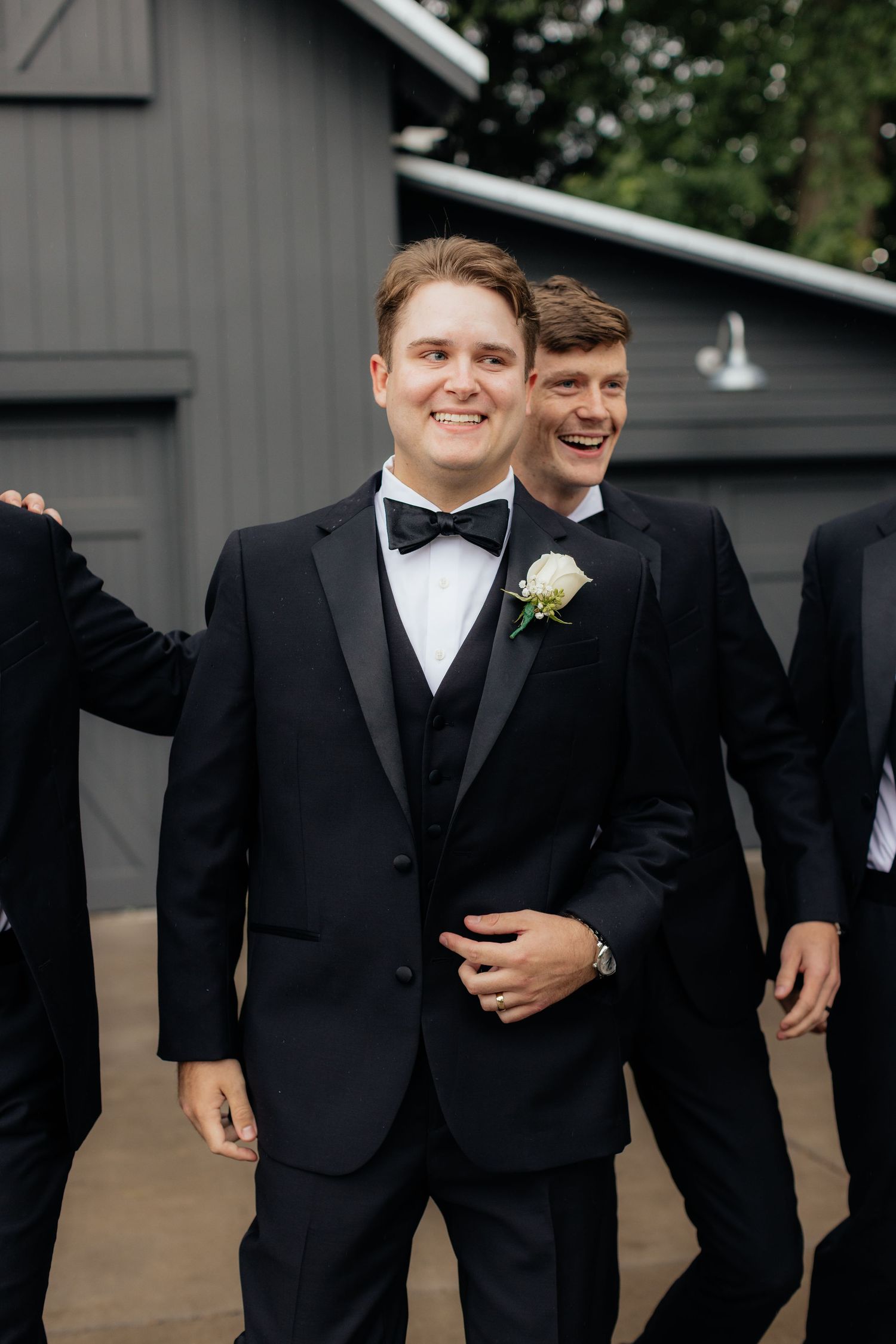 Groomsmen in black tuxedos share candid laughs during outdoor wedding photos.