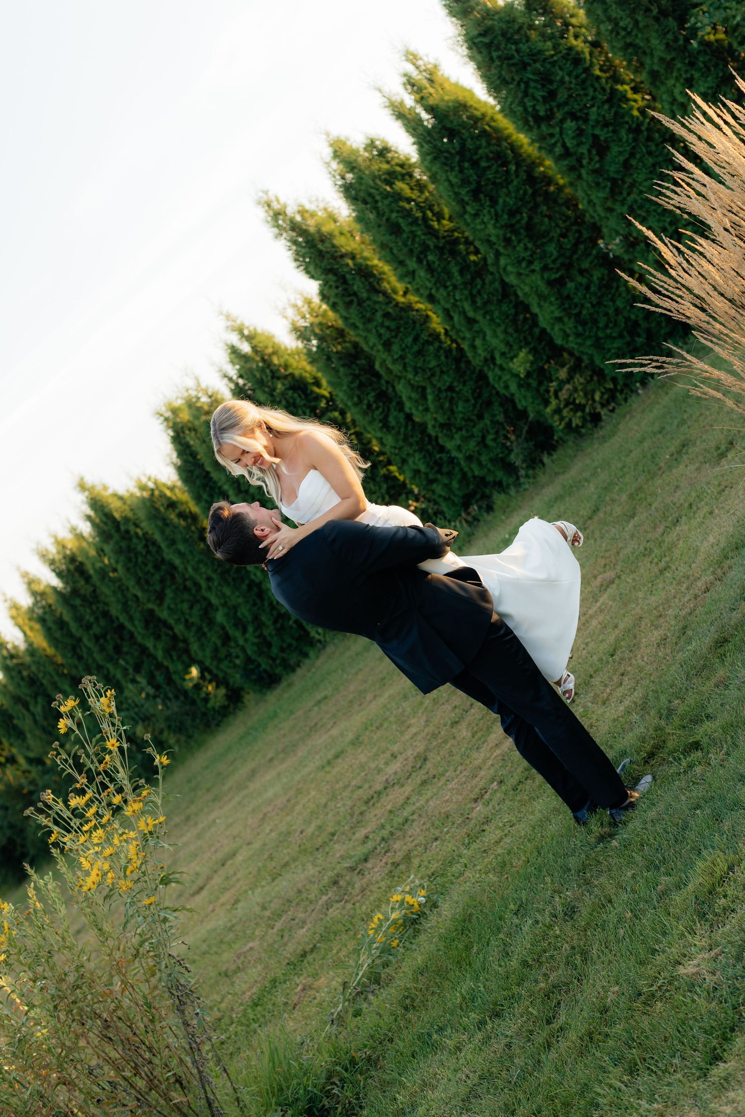 Wedding couple in dramatic pose on grassy hill with evergreen trees at sunset.