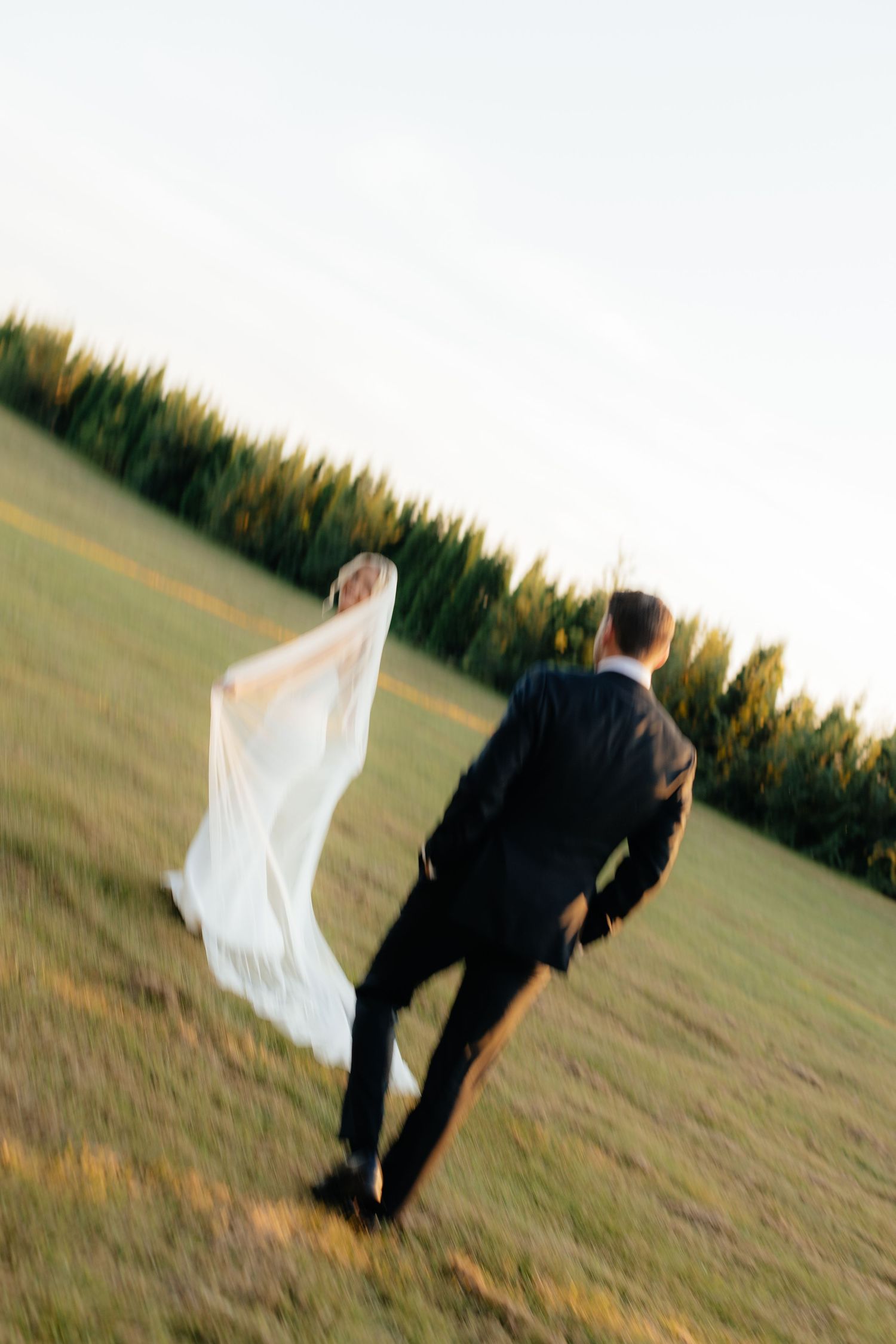 Artistic tilted angle shot of newlyweds walking on grass with evergreen backdrop.