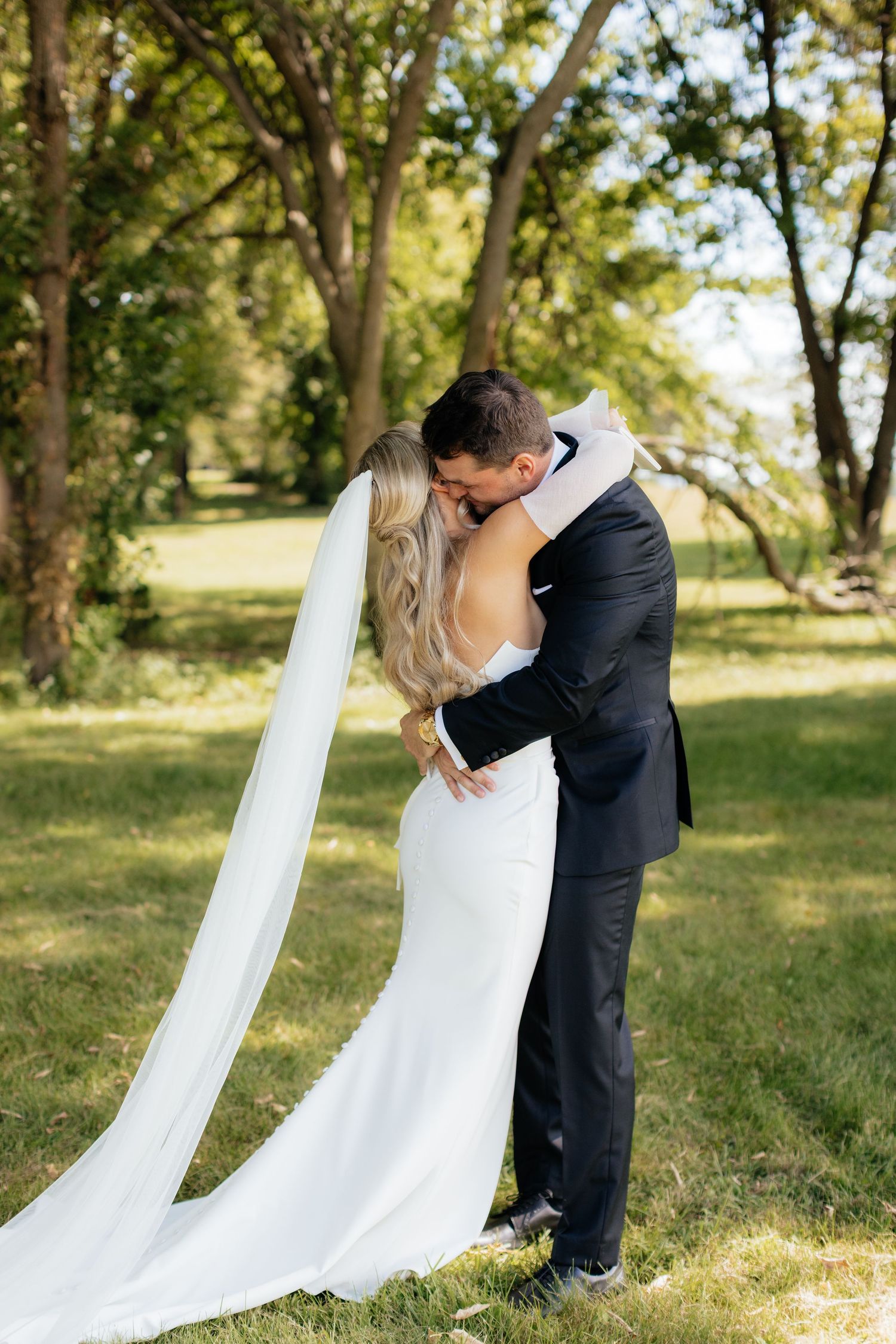 Wedding couple embraces on grassy hill with flowing white veil in summer setting.