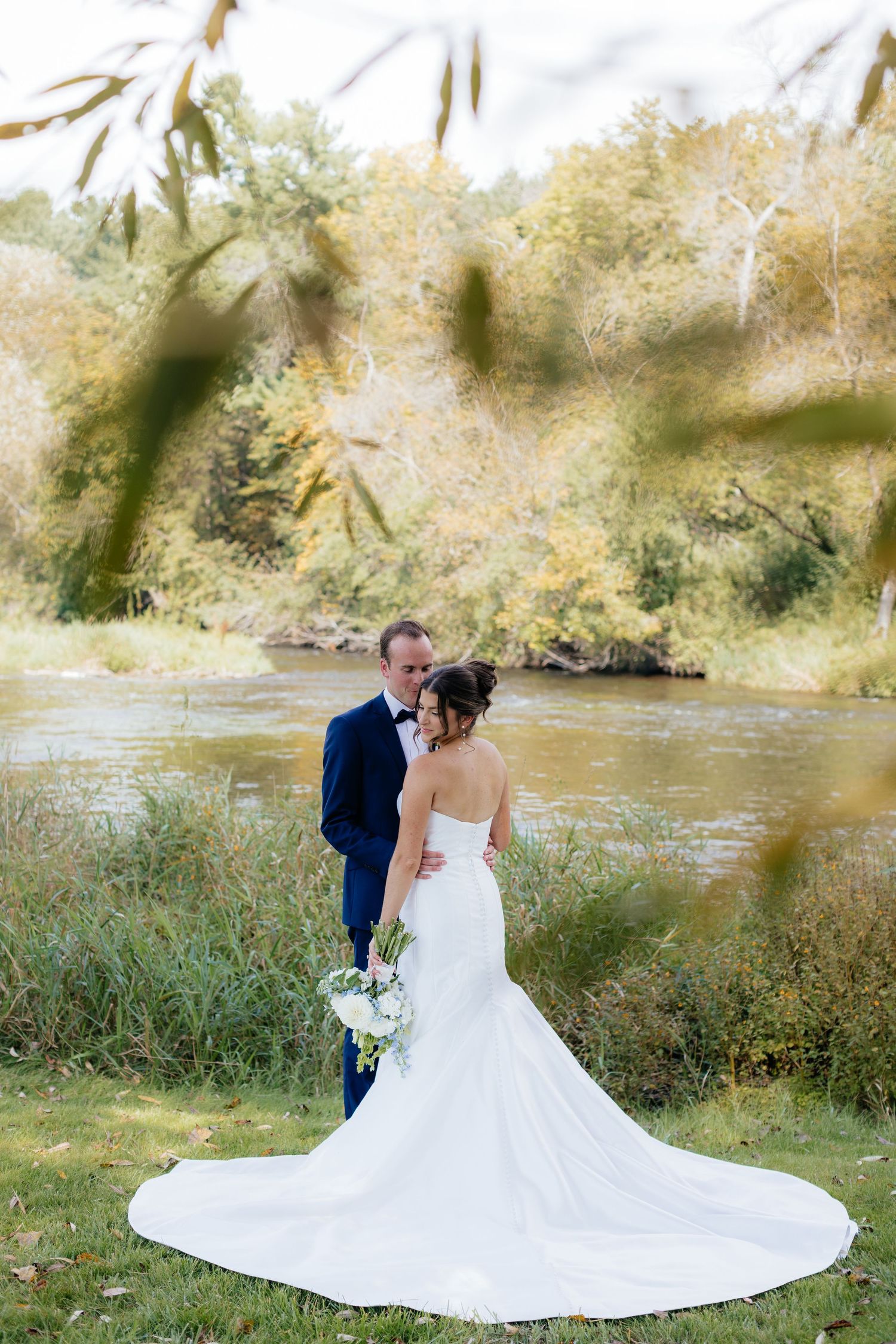 Wedding couple poses by riverside with flowing white dress and natural scenery.