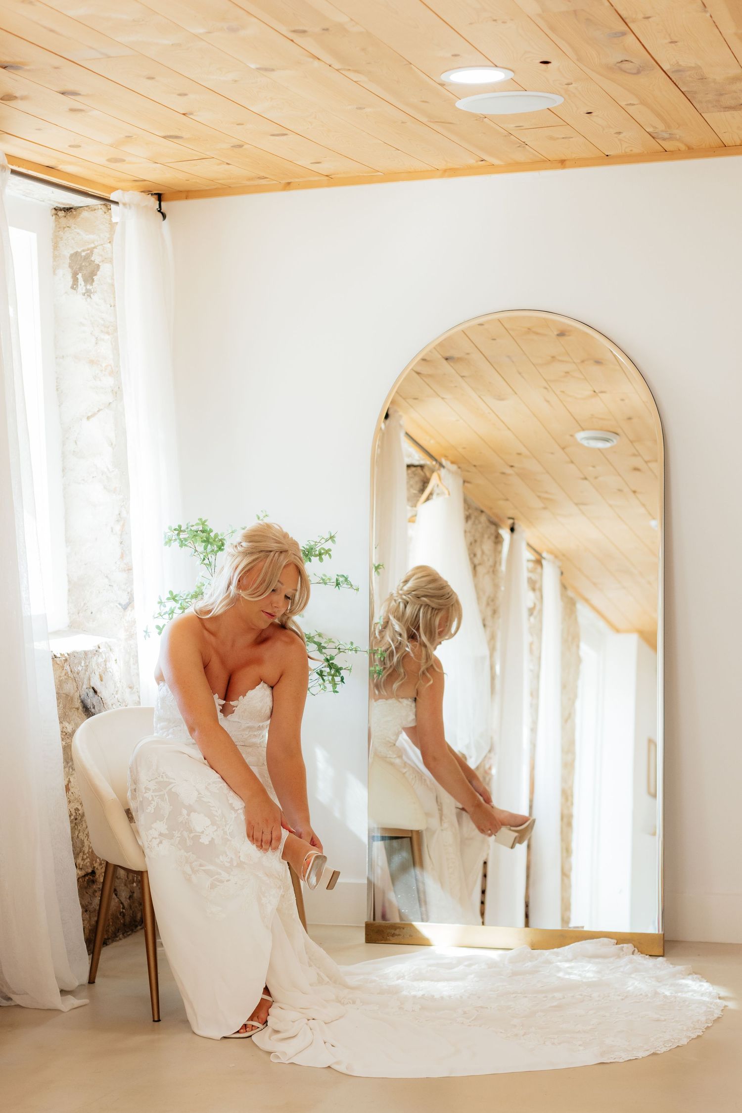 Bride in white wedding dress looks at reflection in full-length mirror in bright room with wood ceiling.