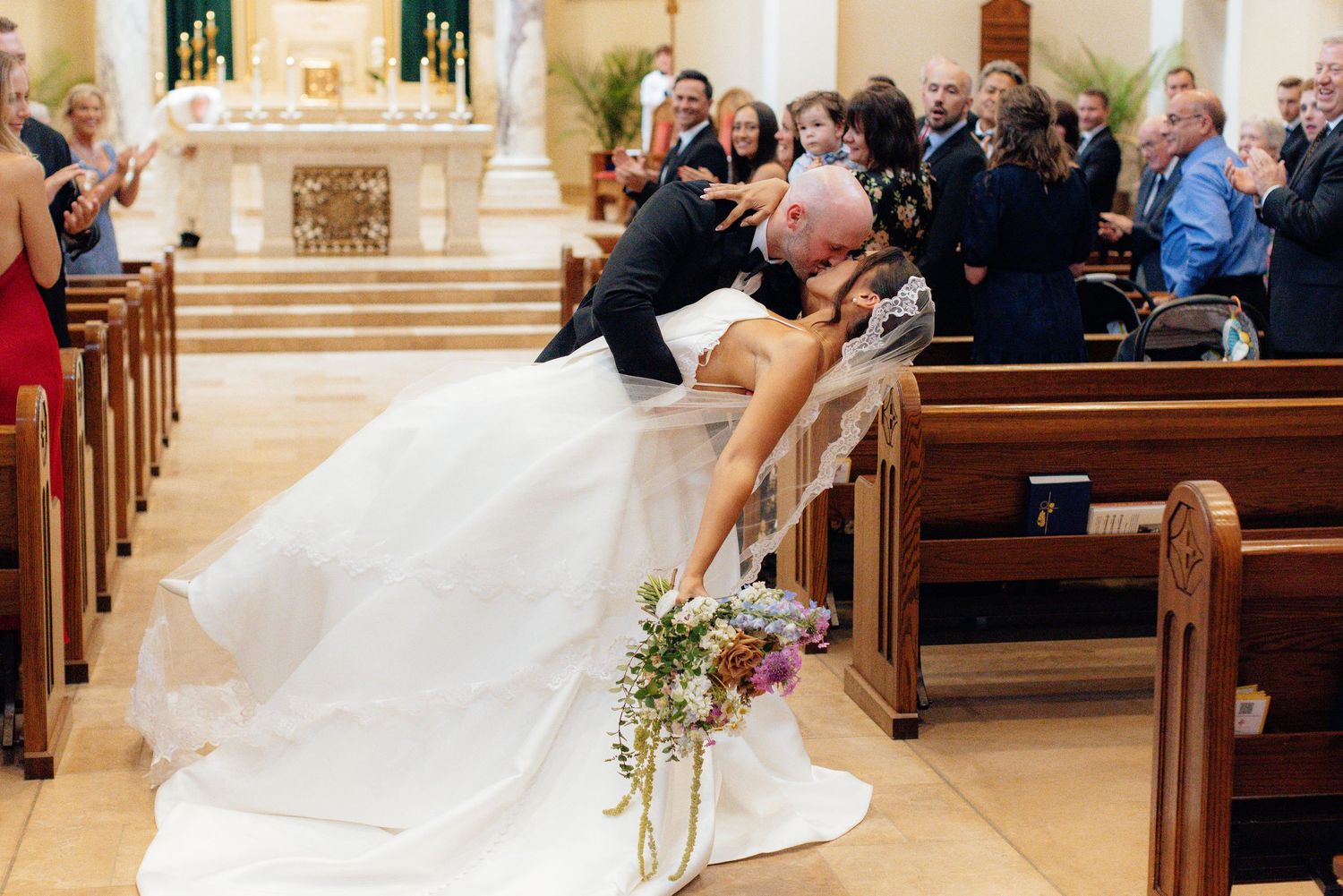 Wide view of wedding ceremony in ornate church with guests filling wooden pews.