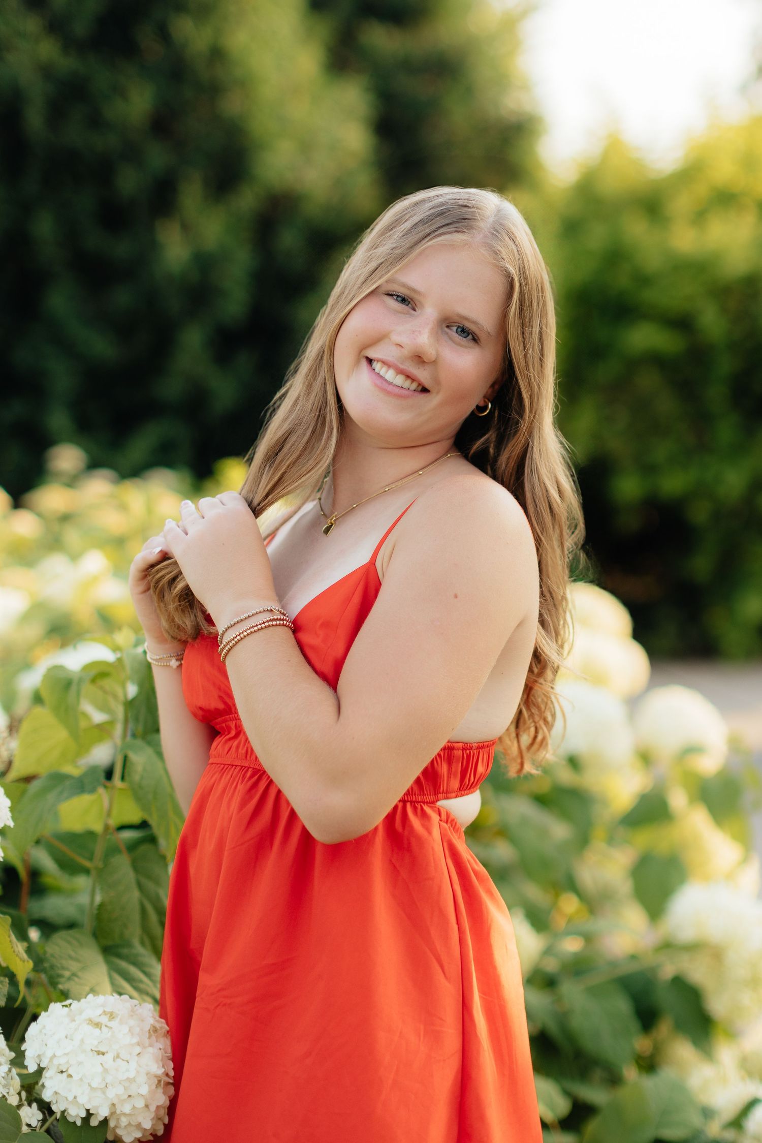 A bright coral sundress is worn in a garden setting with white hydrangea flowers.