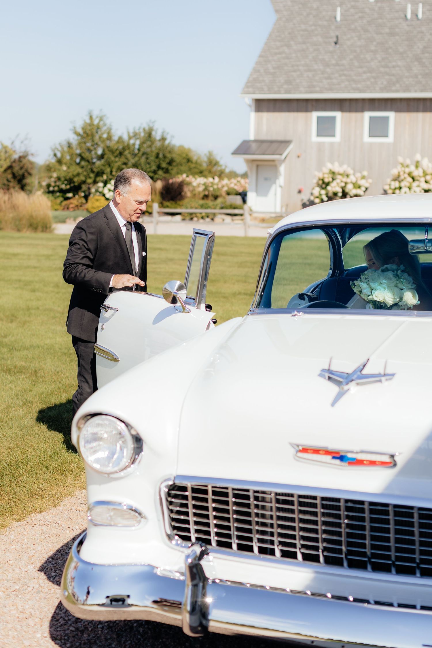 Vintage white Chevrolet parked on lawn near rustic barn building.