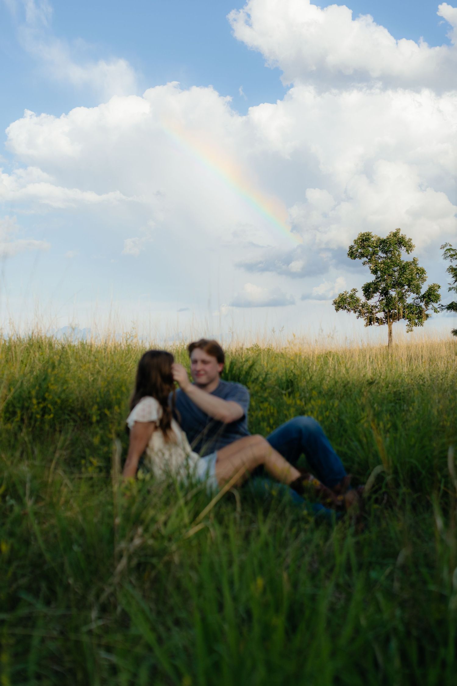 Couple lying in tall grass under blue sky with puffy clouds and rainbow.