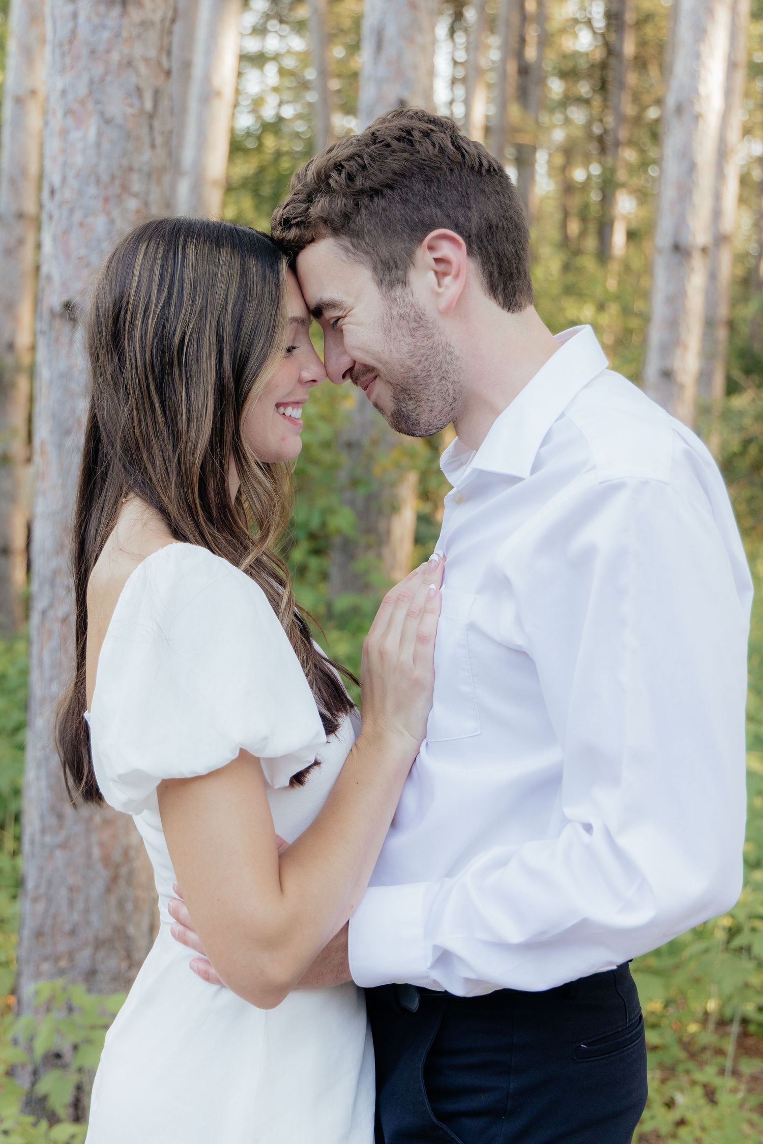 A tender moment between a couple in white attire among tall forest trees.
