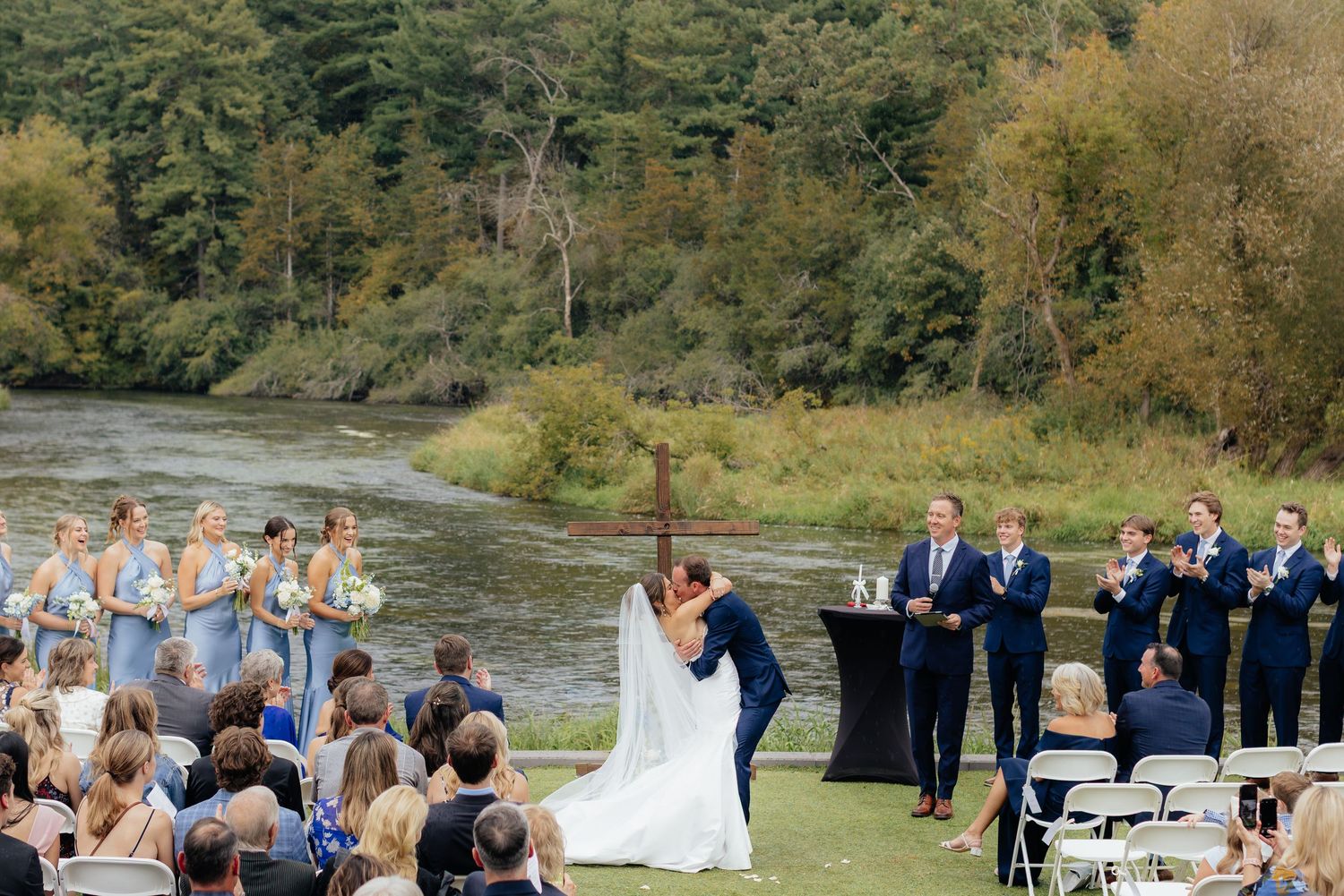 Wedding guests seated outdoors at a ceremony with white chairs and forest backdrop.