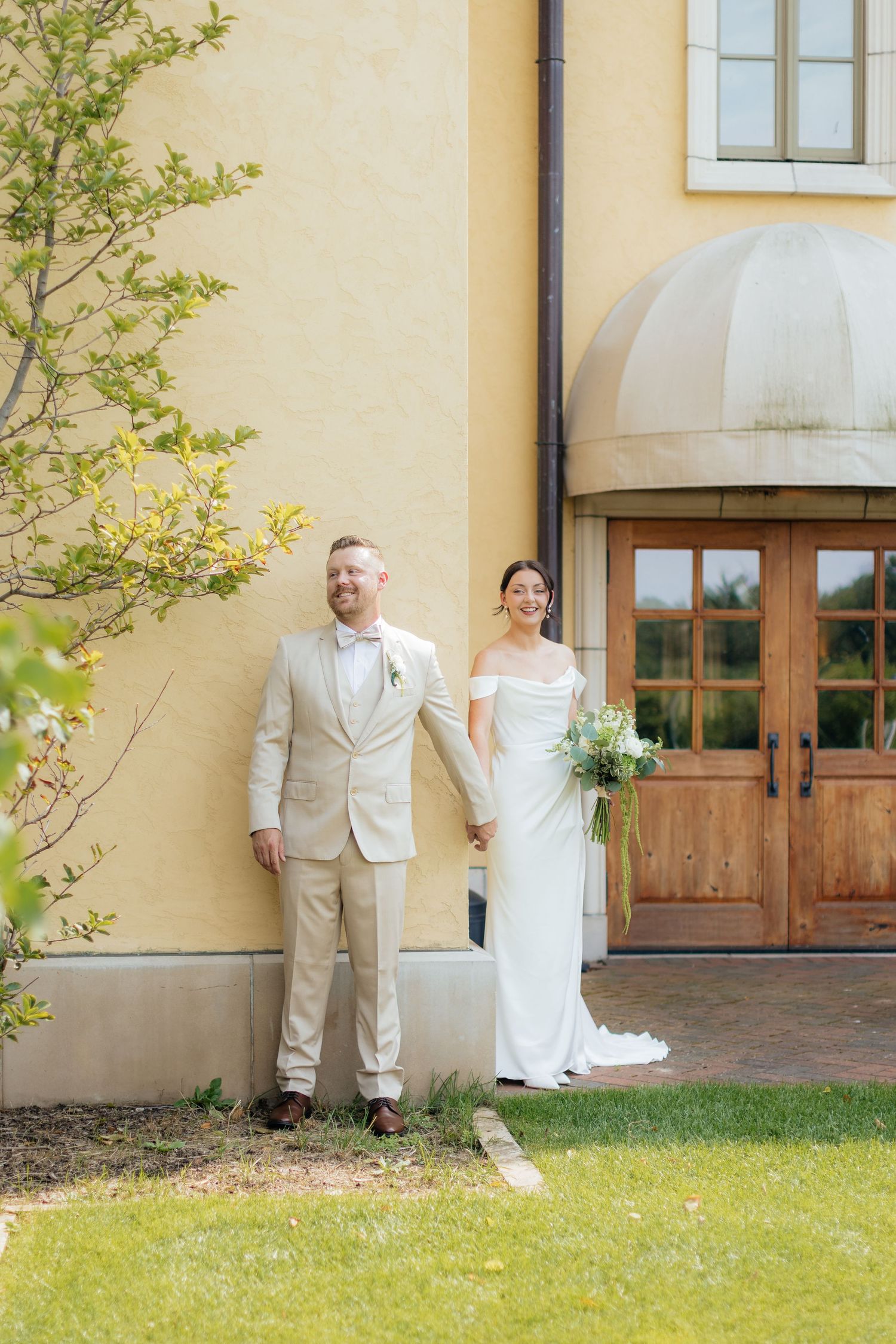 Wedding couple in white dress and beige suit pose outside a yellow stucco building.