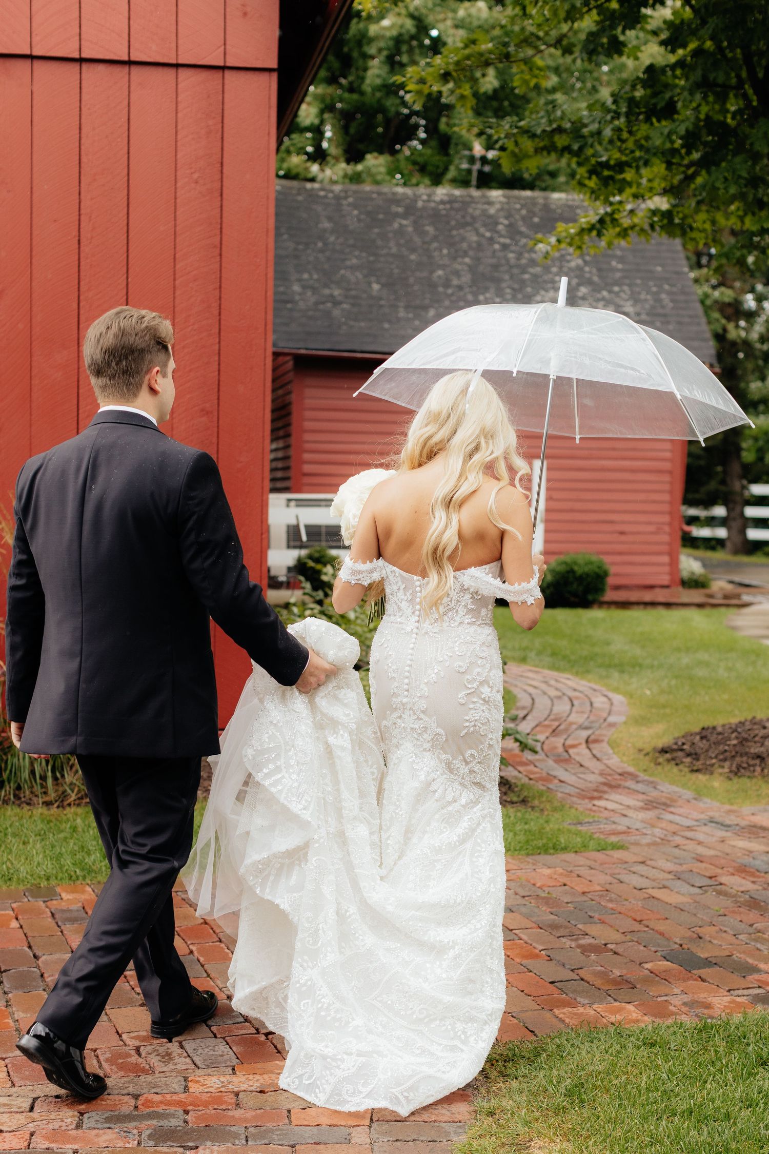 Bride and groom stroll past red barn on brick pathway during wedding photoshoot.