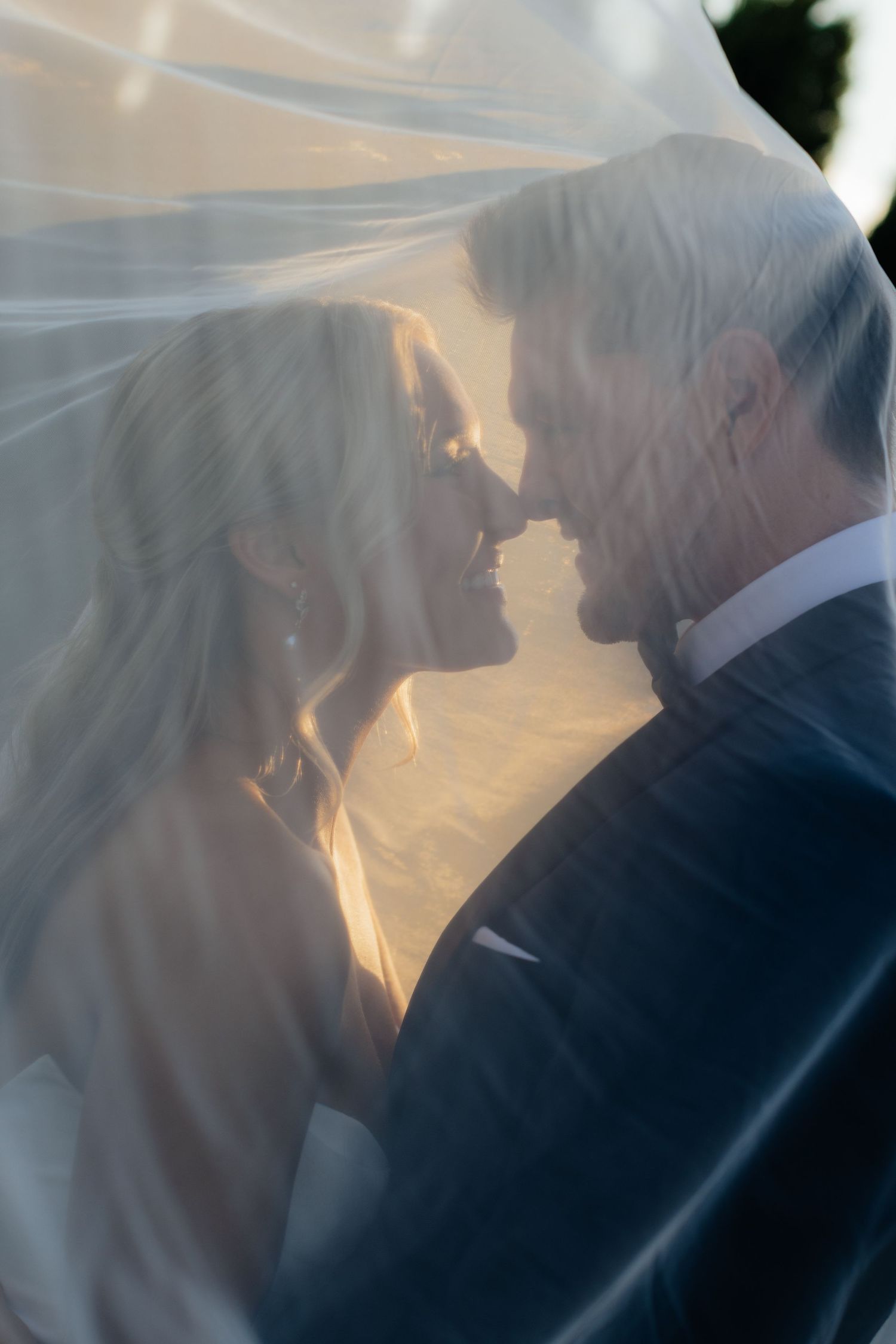 A romantic wedding couple shares an intimate moment under a veil during golden hour.