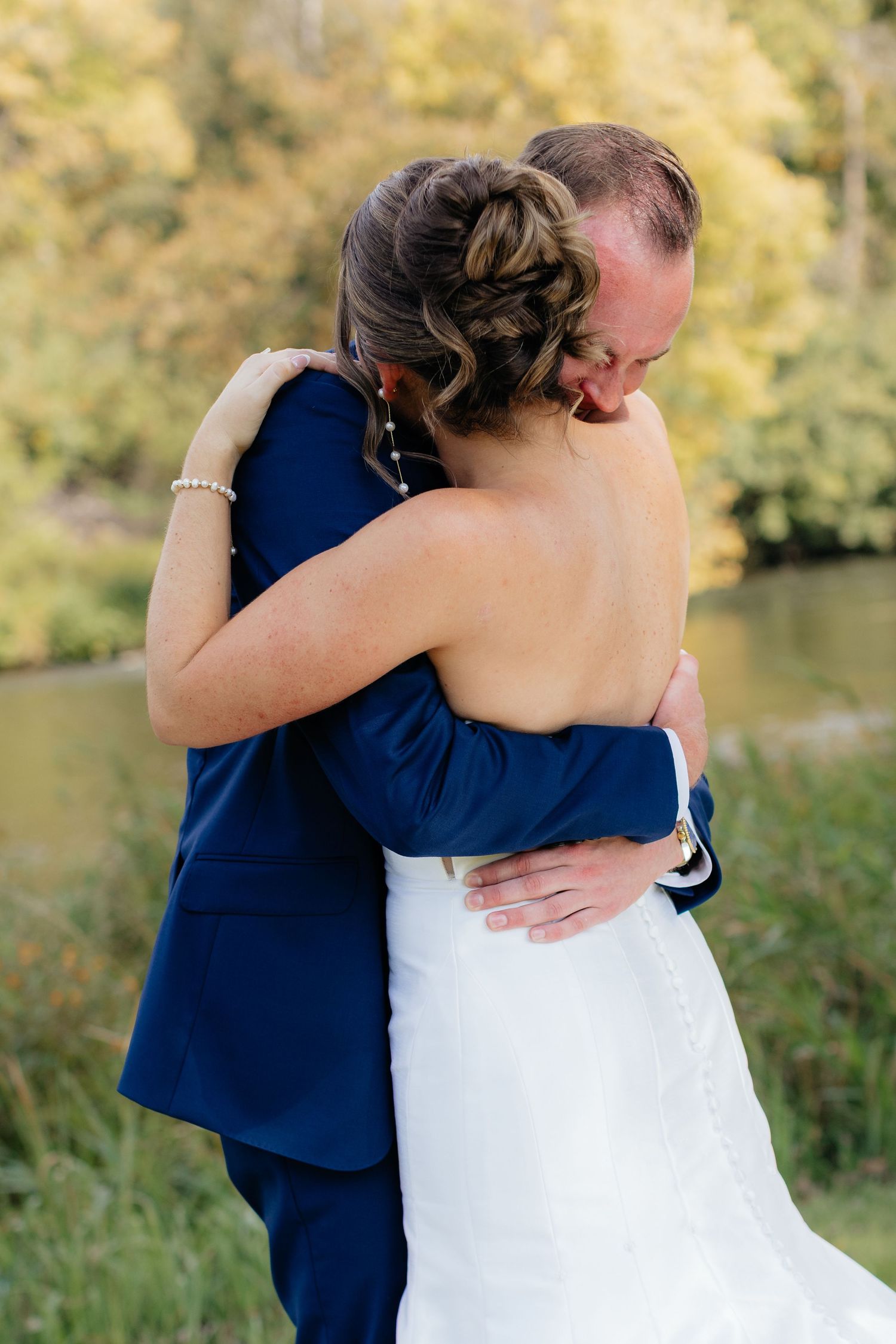 Newlyweds embrace outdoors with autumn foliage in the background.