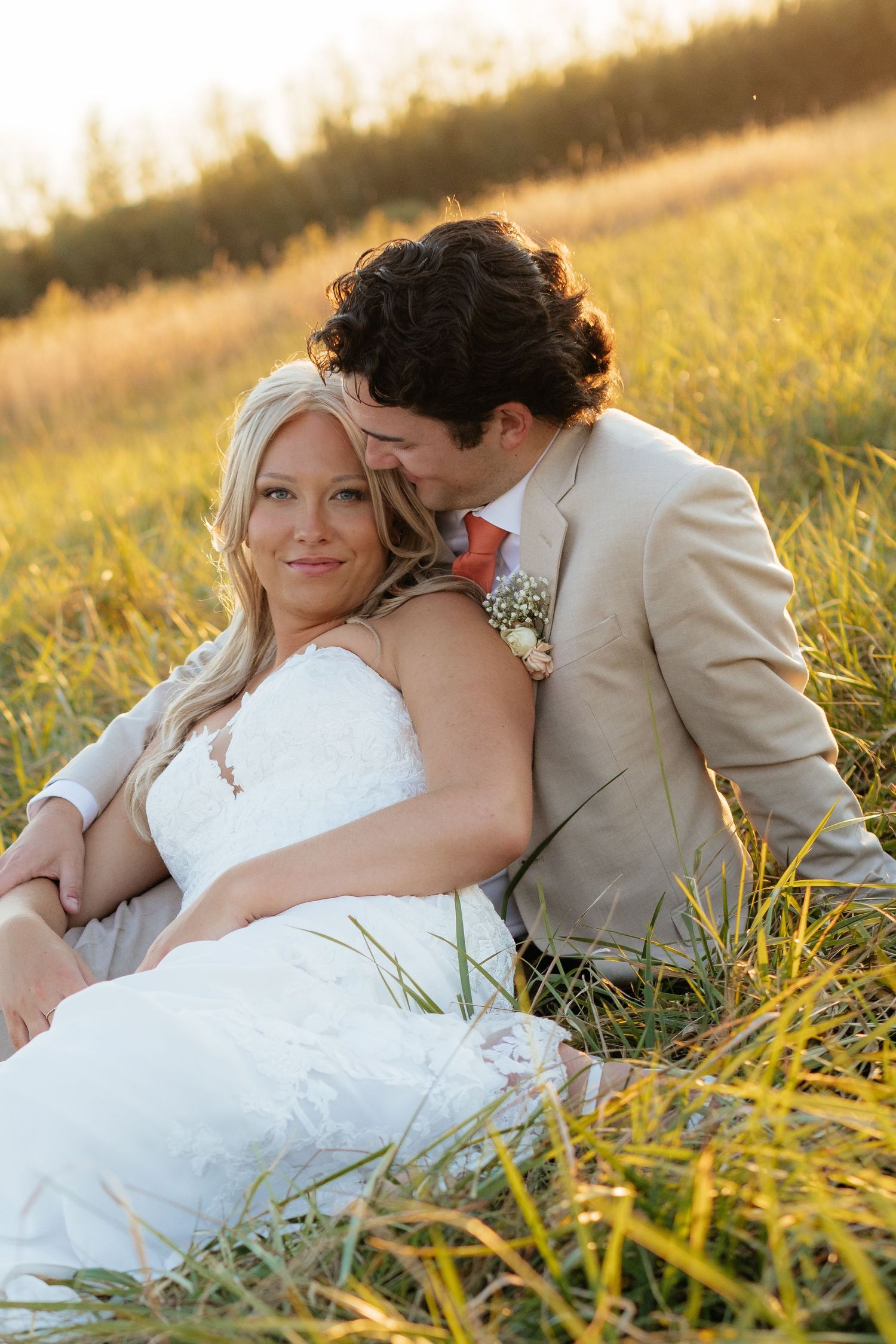 Bride in white dress and groom in tan suit resting together in sunlit meadow.