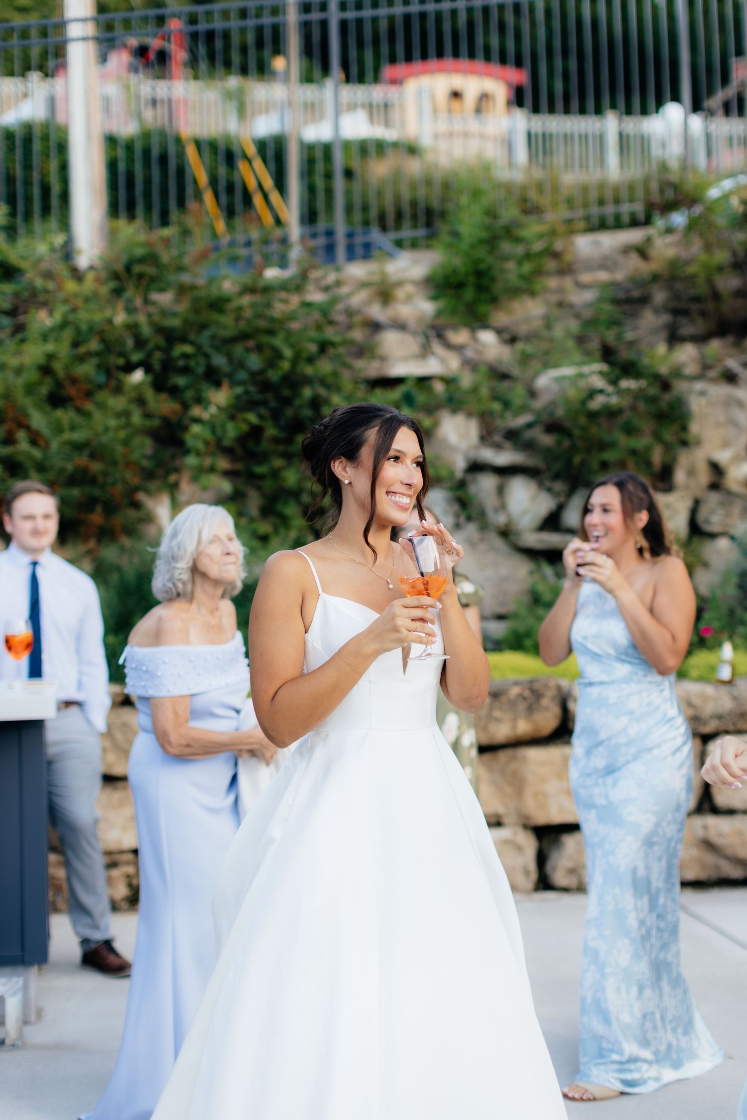 Wedding party celebrates outdoors with drinks and laughter near a stone wall.