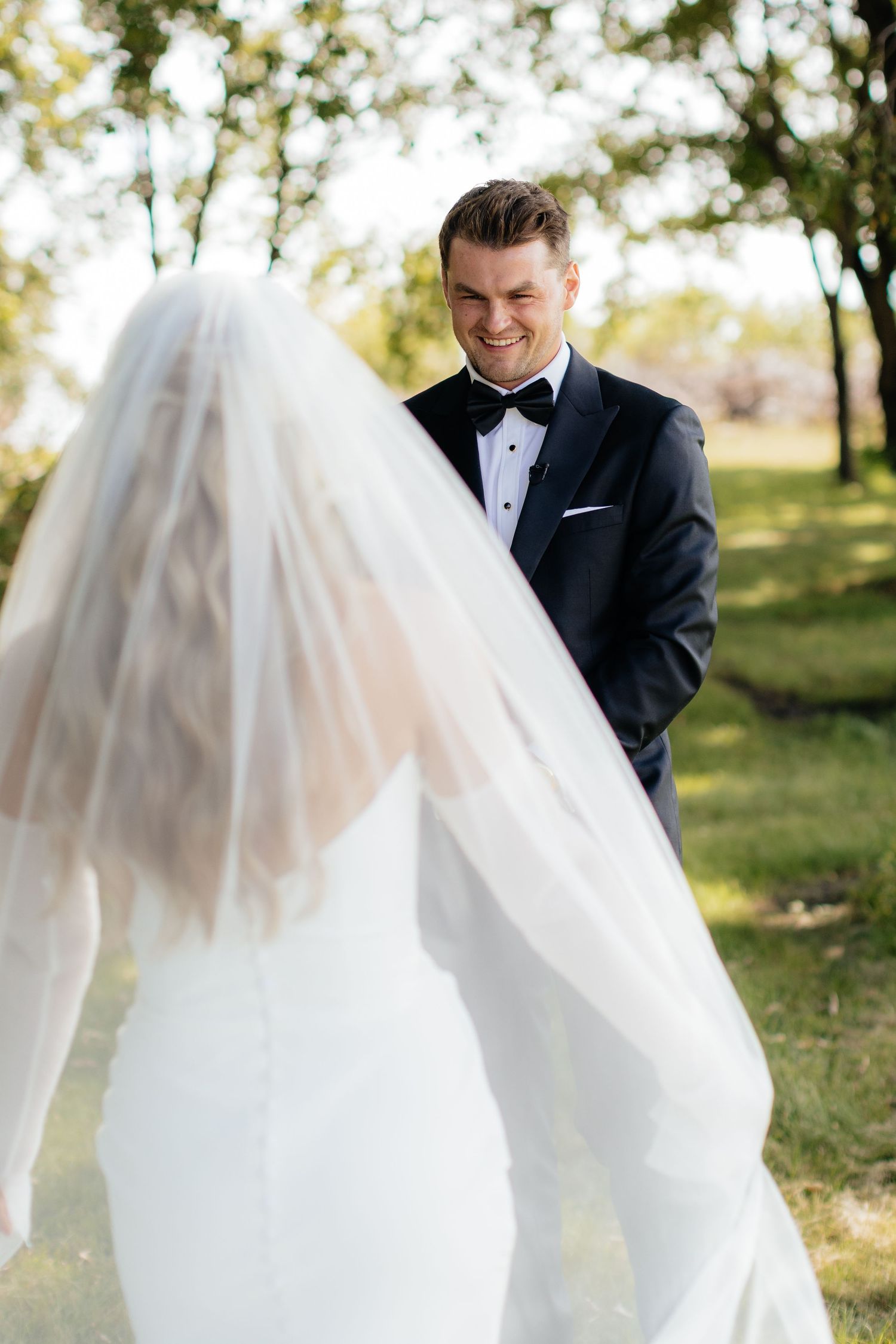 Groom admires bride's flowing veil during outdoor wedding portraits.