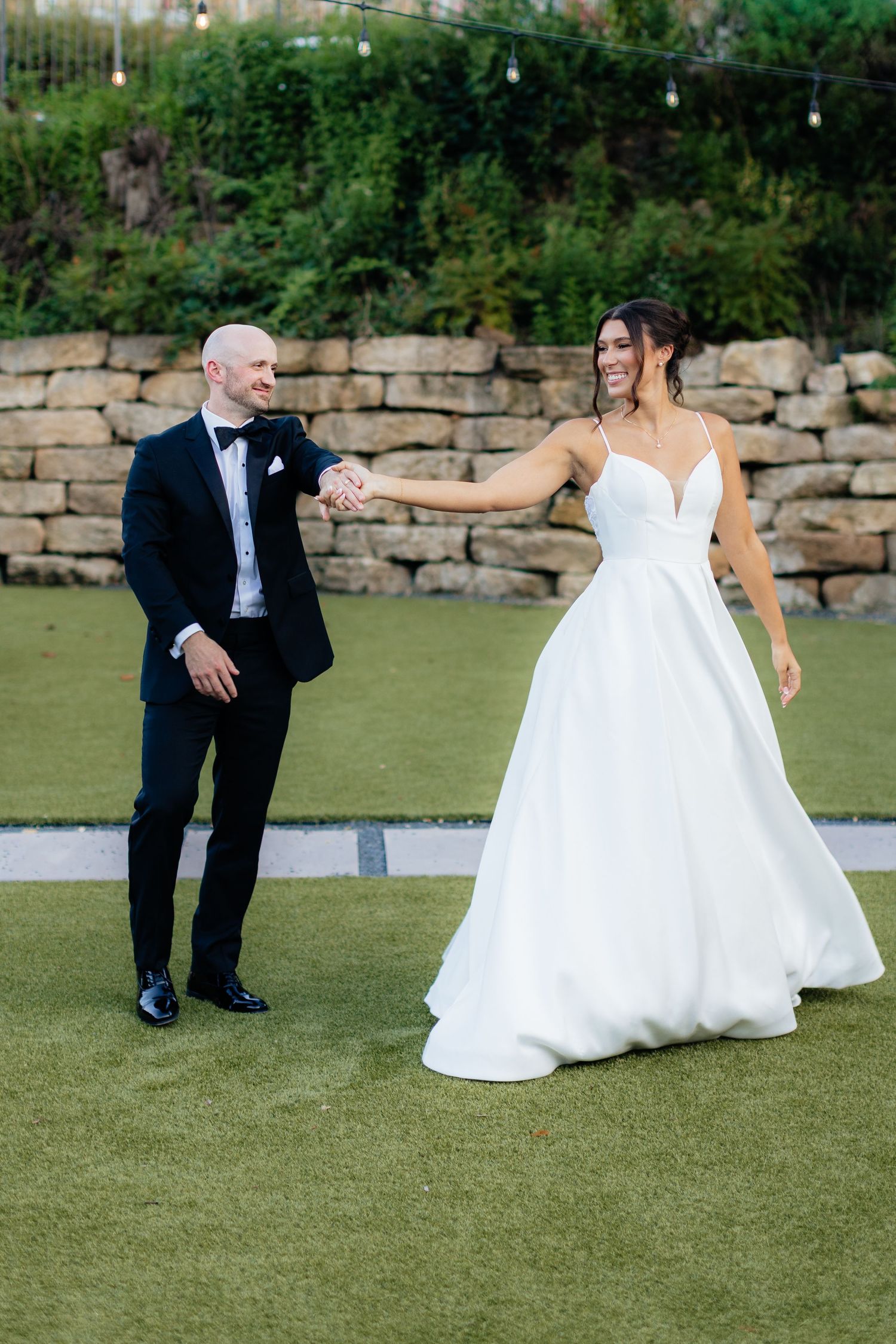 Wedding couple interacts playfully on manicured lawn with stone wall backdrop.