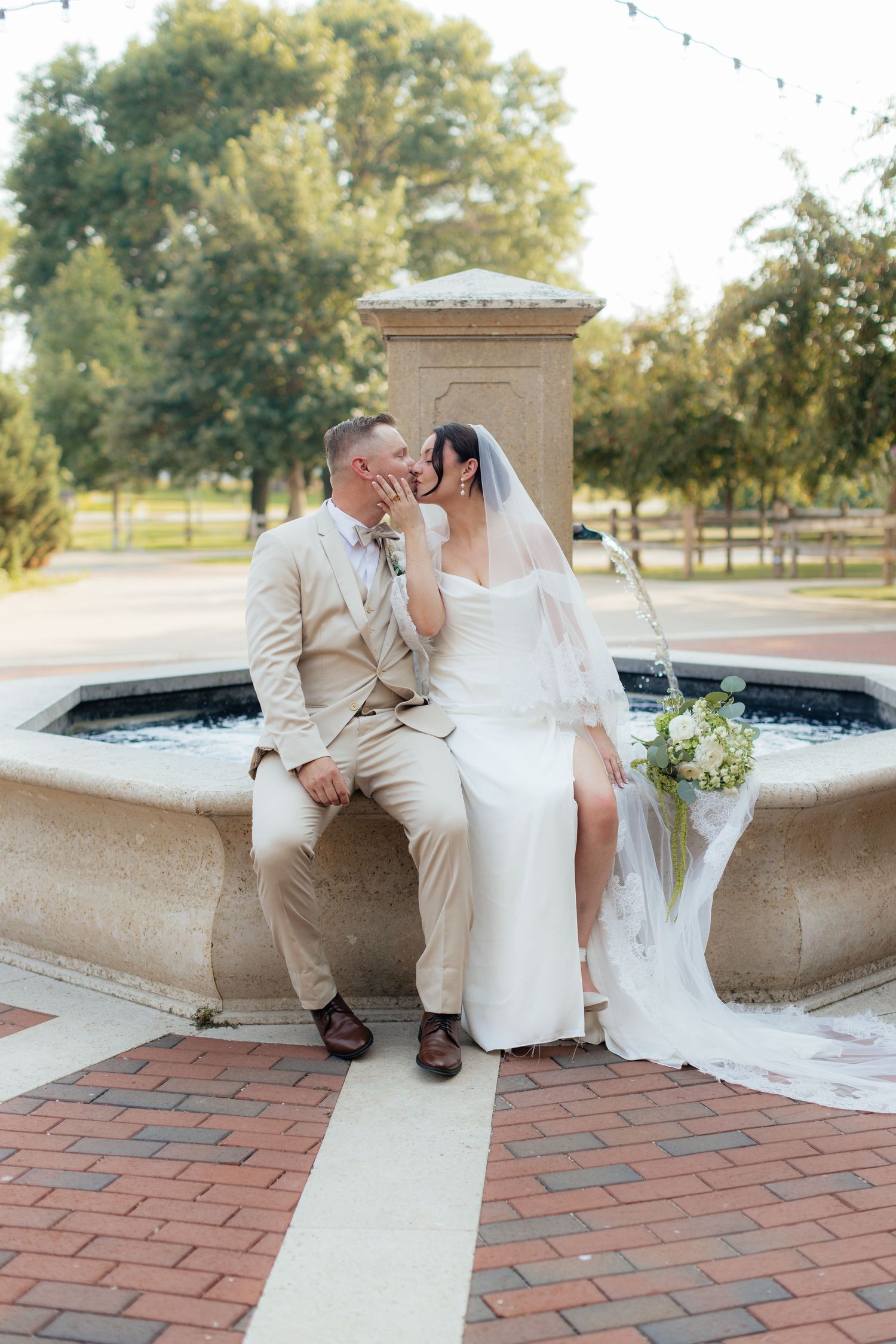 Newlyweds share an intimate moment while sitting on a stone fountain in a garden setting.