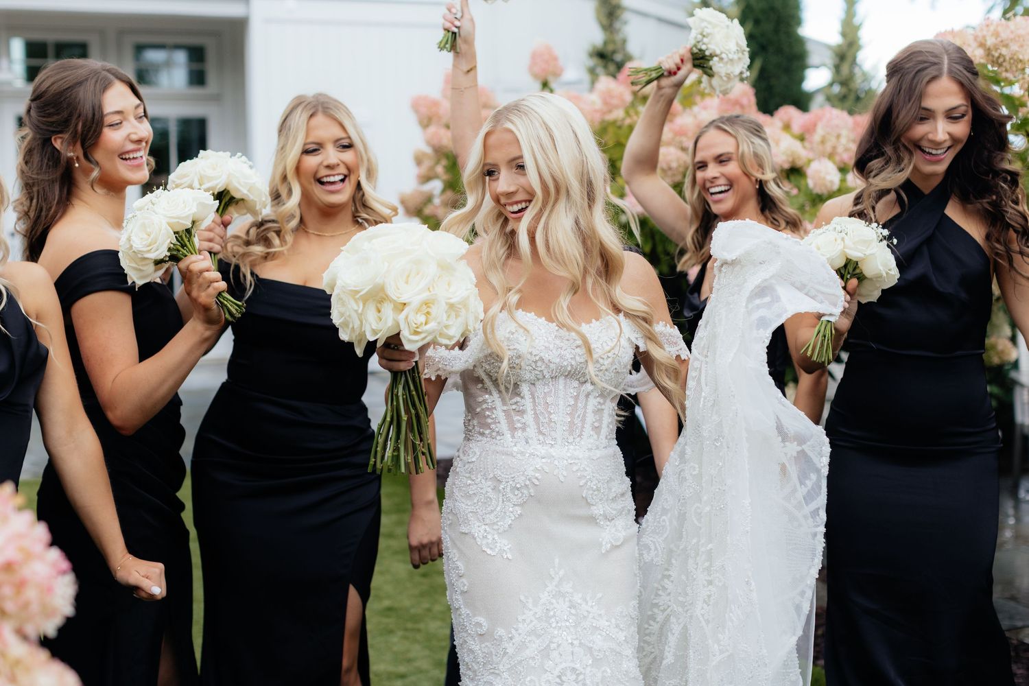 Bridal party in black dresses poses with bride in white lace gown holding white rose bouquets.