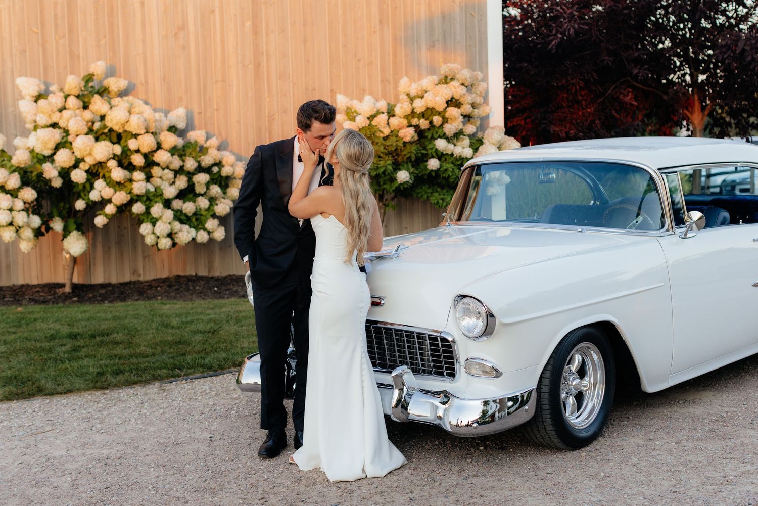 Newlyweds share an intimate moment next to a classic white Chevrolet with peach roses in background.