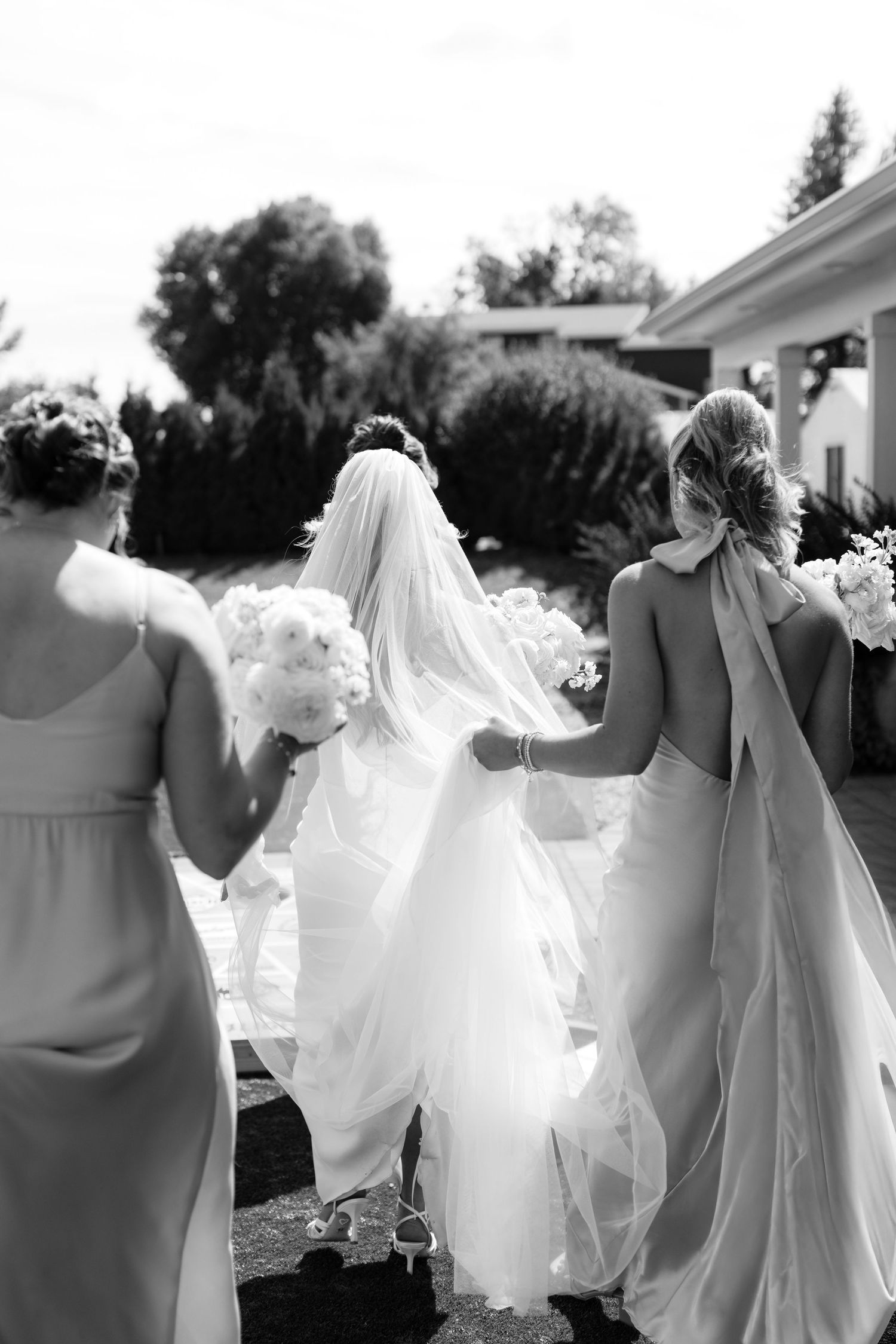 Bridesmaids in elegant dresses assist with the bride's flowing veil during an outdoor wedding ceremony.