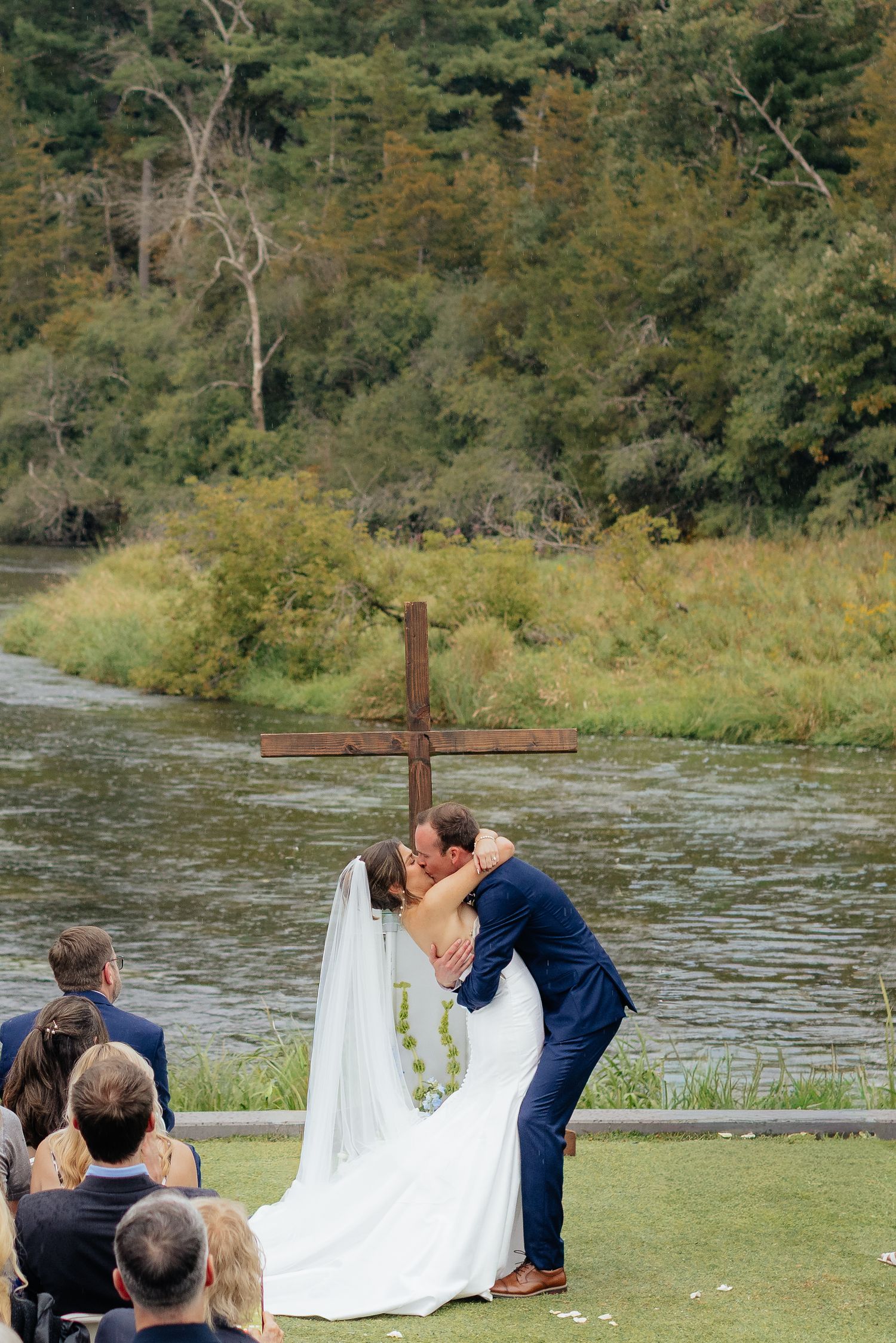 Wedding ceremony taking place by a scenic river with wooden cross backdrop and seated guests.