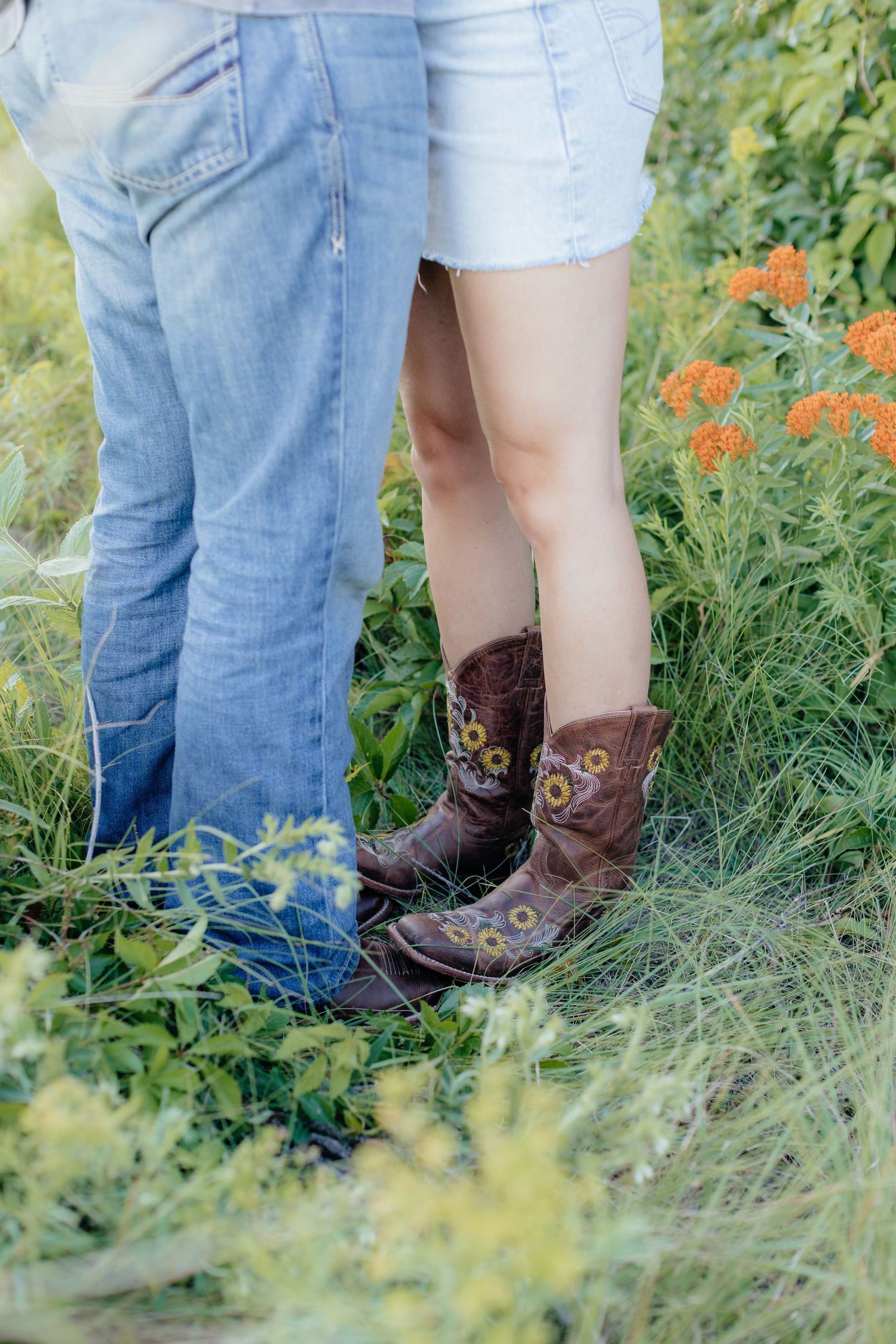 Close-up of cowboy boots worn by two people standing in grass with orange wildflowers.