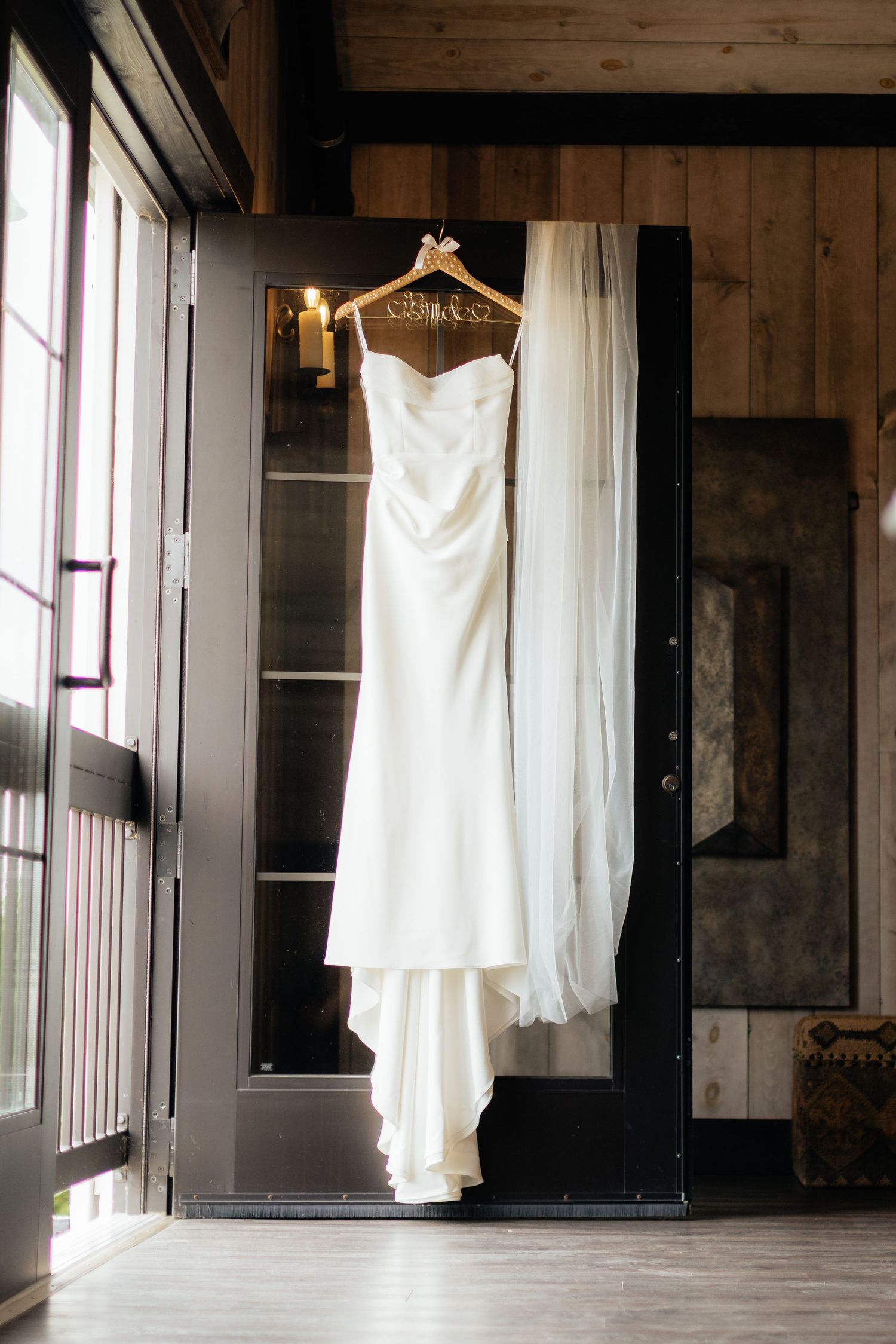 A classic white wedding dress hanging on a wooden hanger in a rustic room.