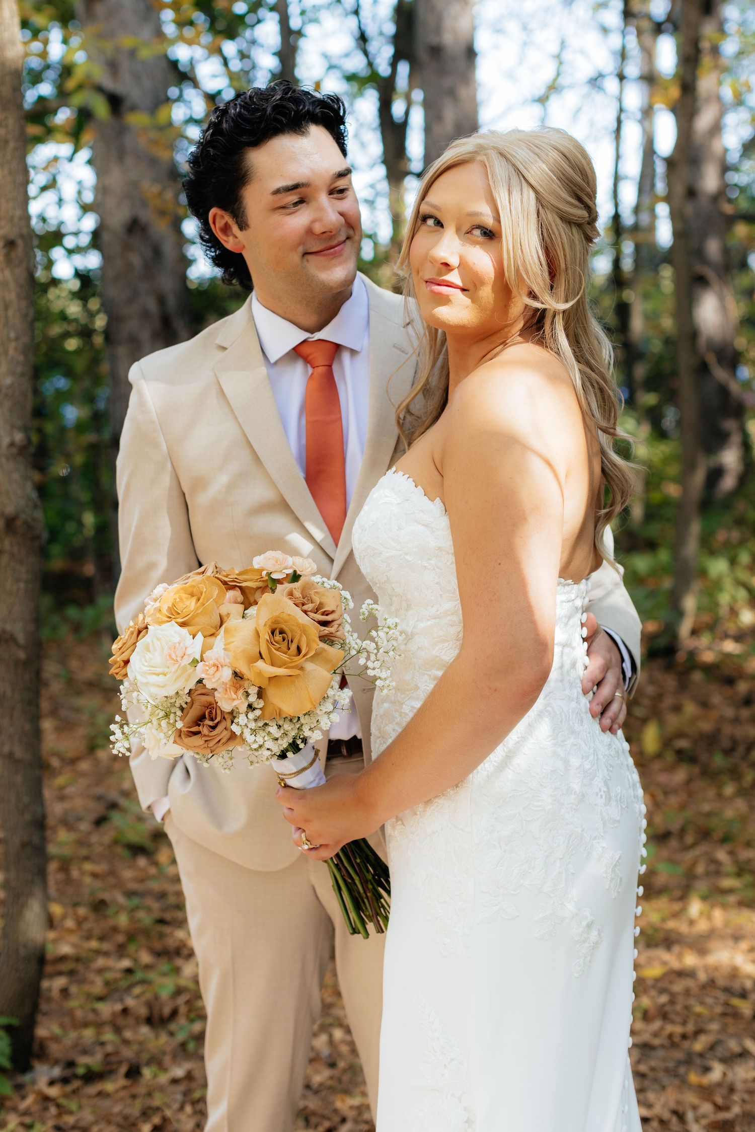 Wedding couple poses with peach and cream bouquet in wooded setting.