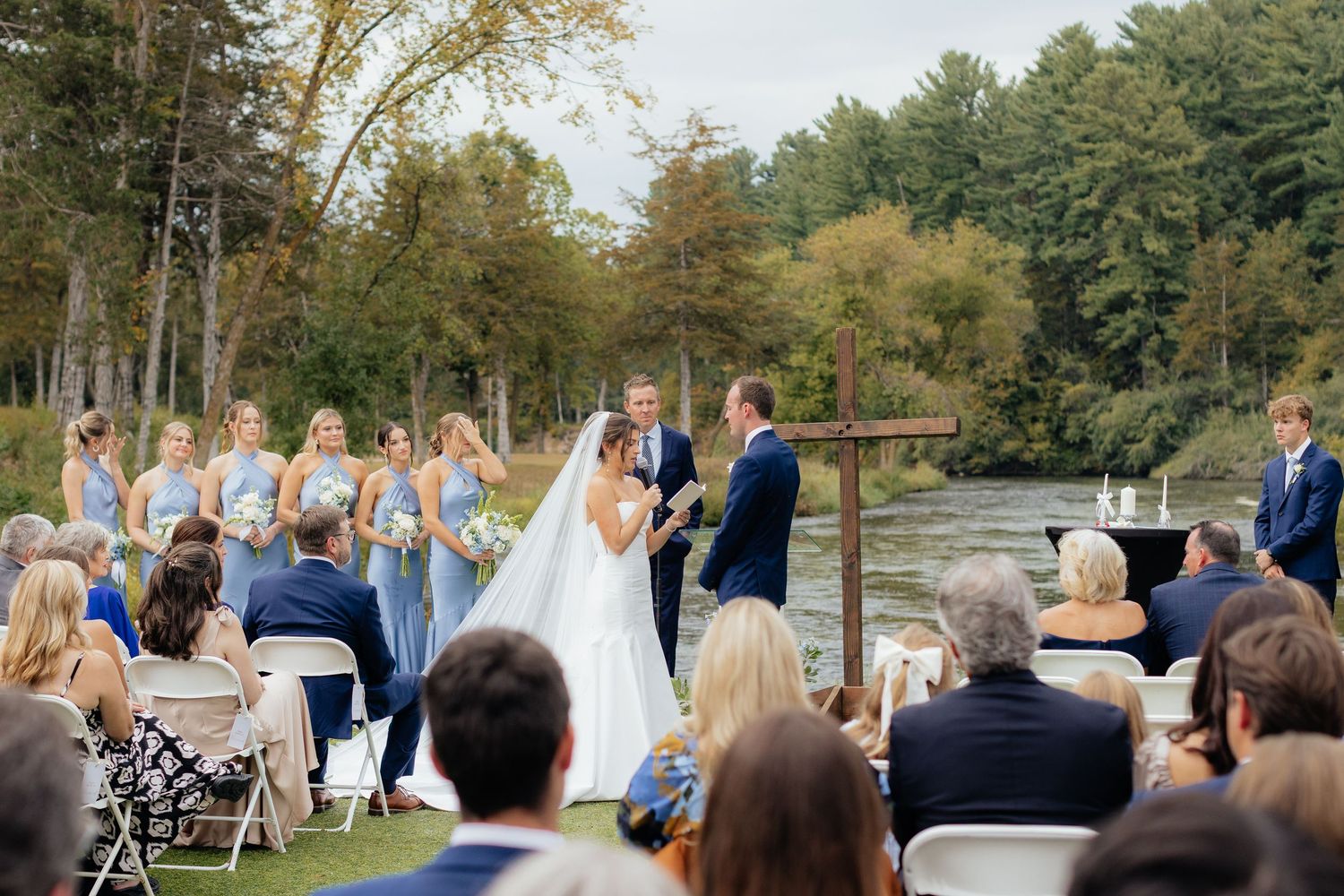 Outdoor riverside wedding ceremony with wooden cross altar and seated guests in natural setting.