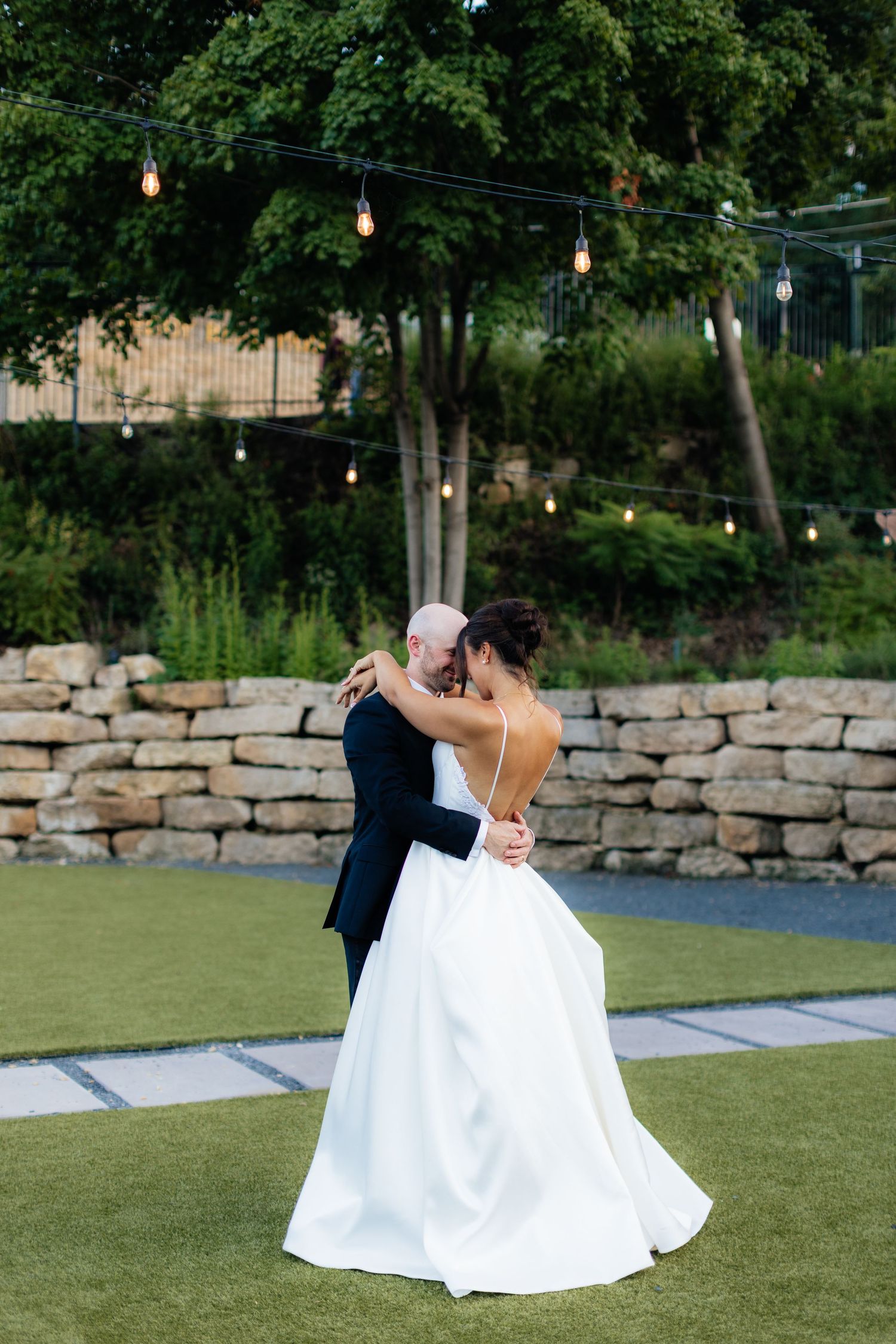 A couple shares an intimate dance moment on a lawn with string lights and stone wall backdrop.