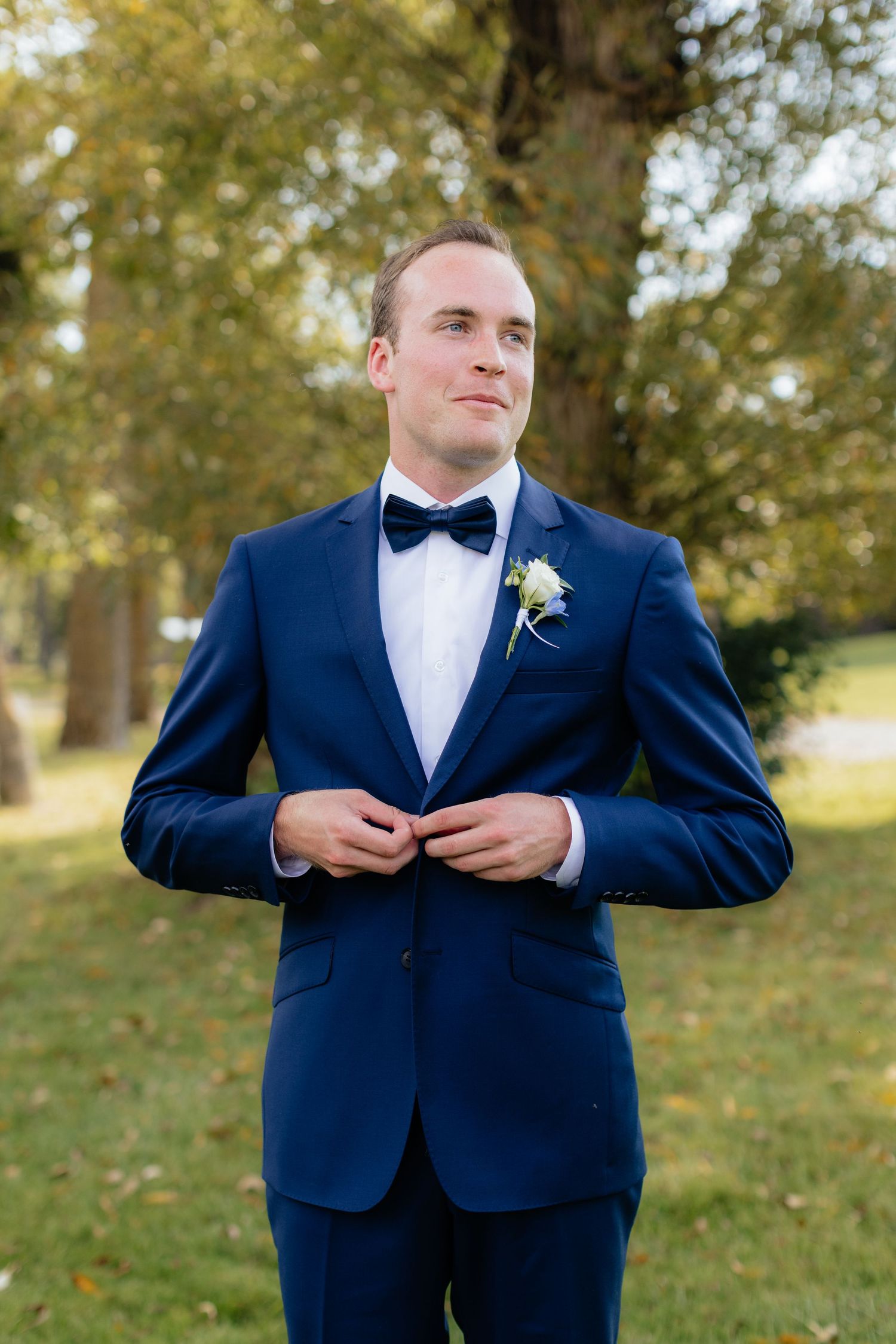 Groom in navy suit and bowtie prepares for wedding ceremony outdoors.