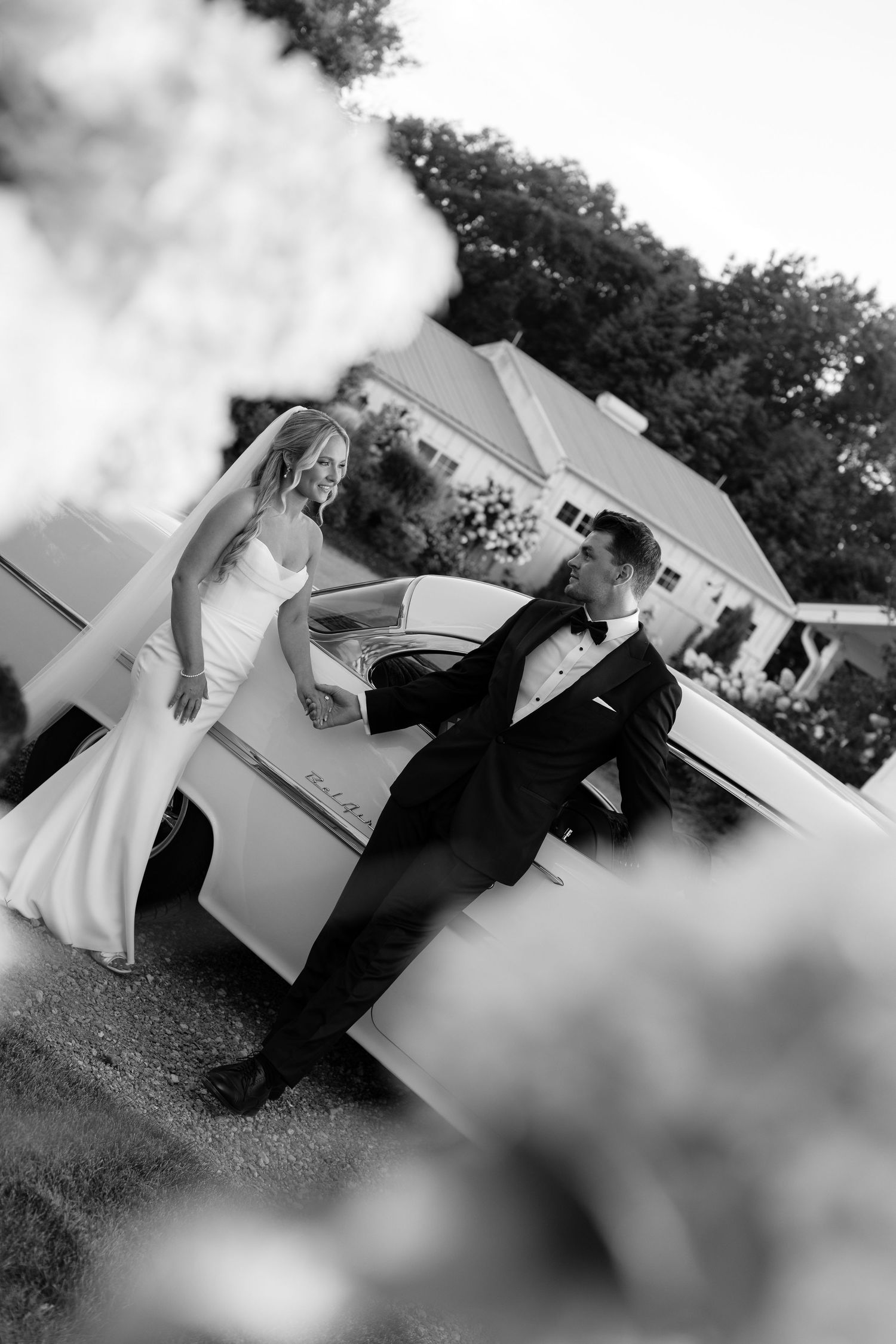 Black and white photo of wedding couple posing playfully by classic car.