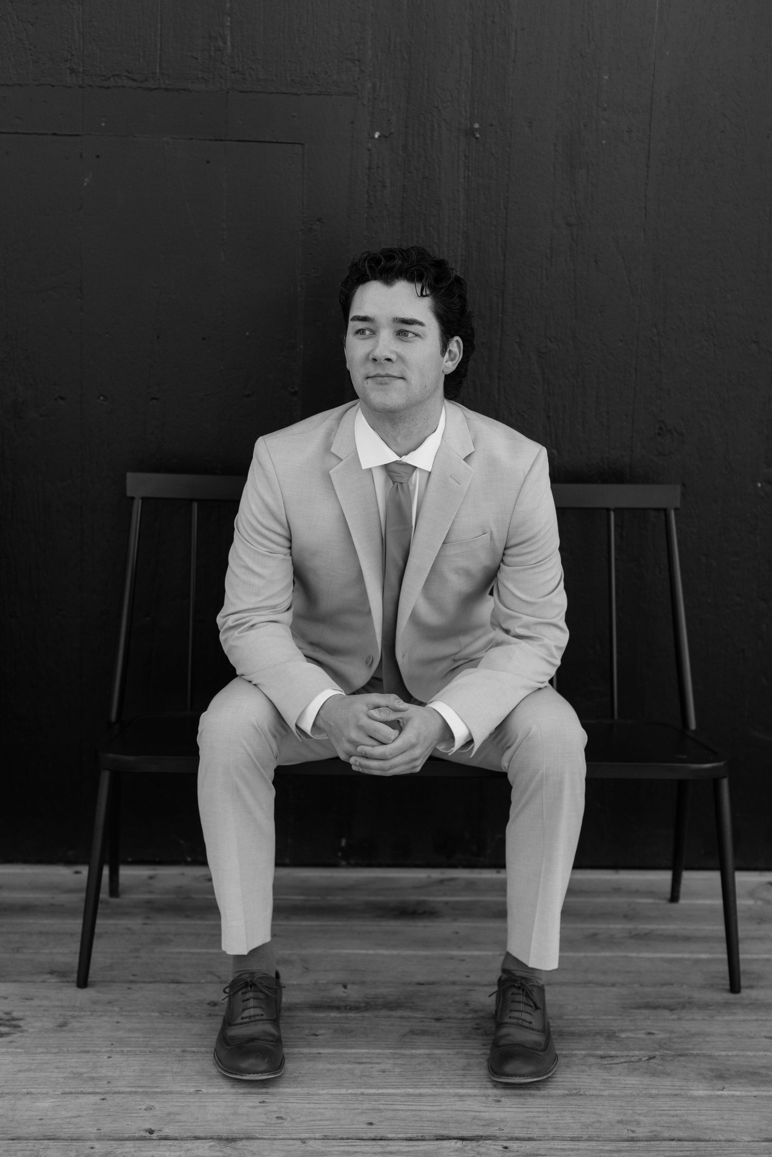 Person in light suit sitting on wooden bench with dark background in artistic portrait series.