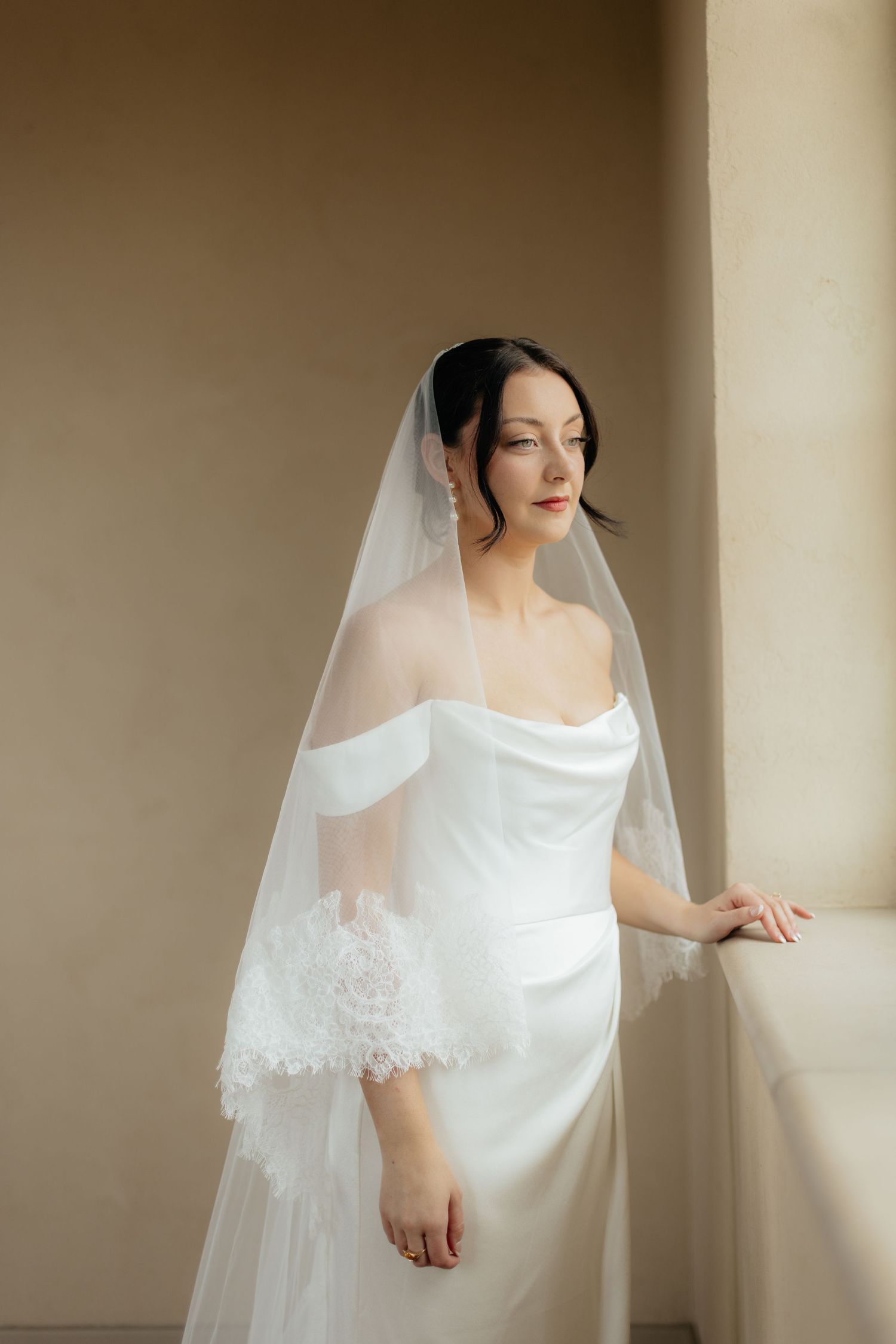 Elegant bride in a simple white wedding dress and veil standing in soft natural light.