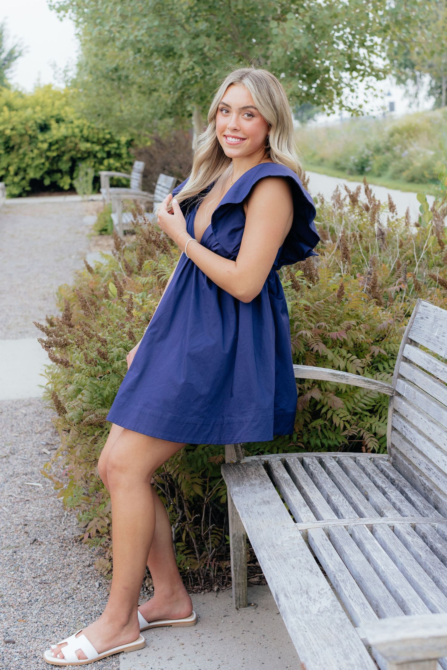 A navy blue sleeveless dress is styled with white sandals in an outdoor garden setting.
