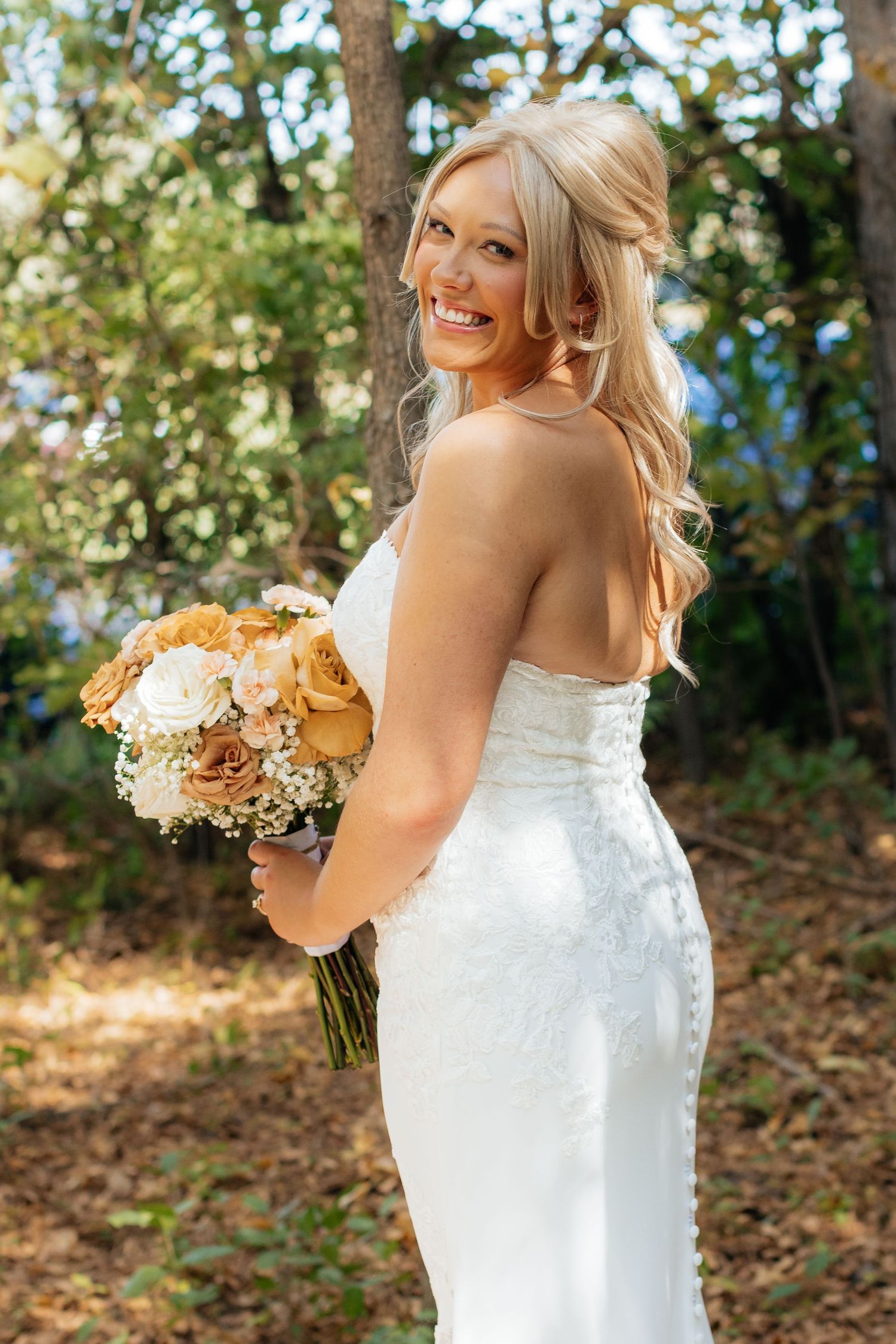 Bride in strapless gown holds peach and cream bouquet while posing in outdoor forest setting.