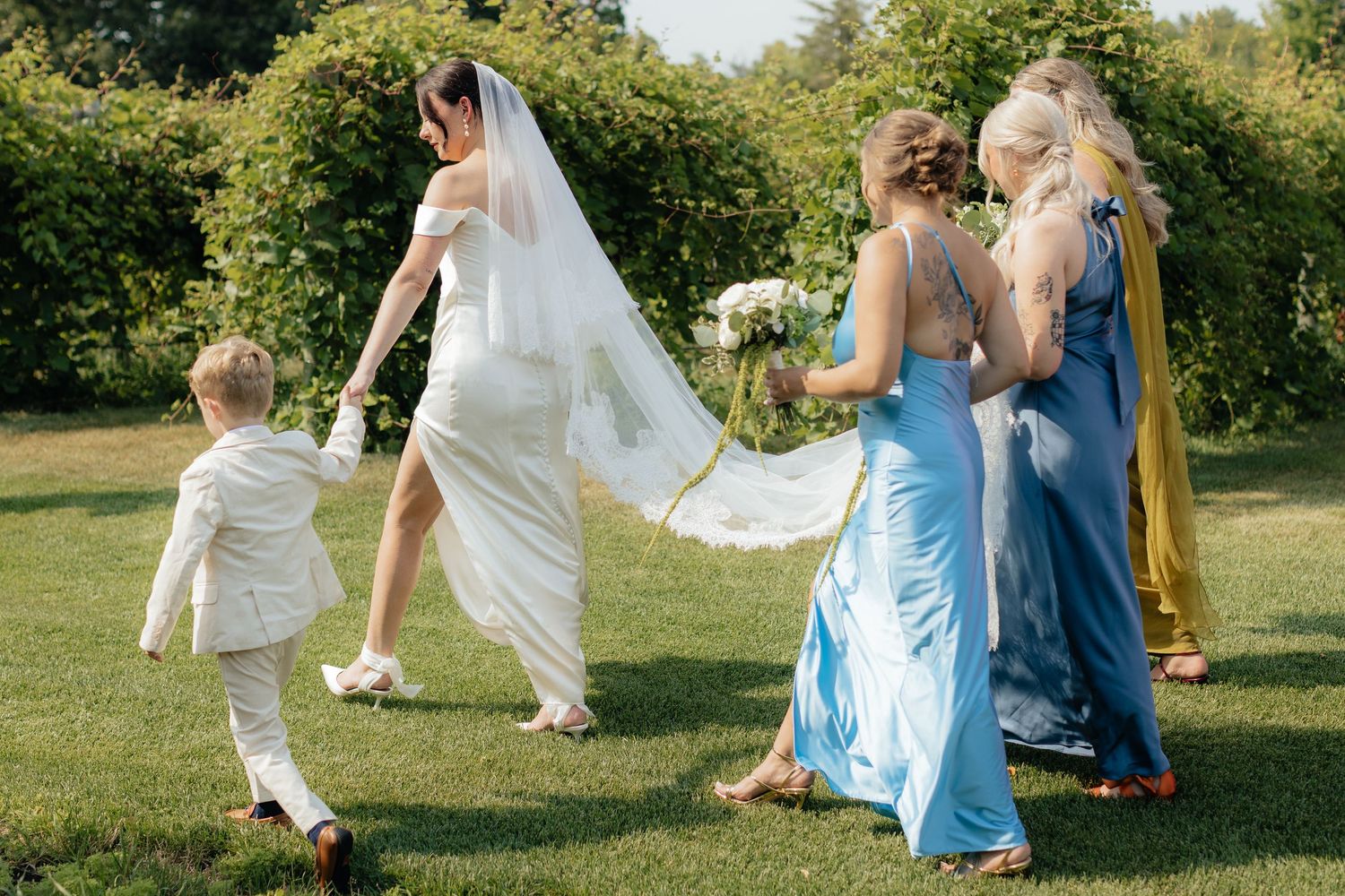 Wedding party members in white and light blue attire dance joyfully on green grass outdoors.