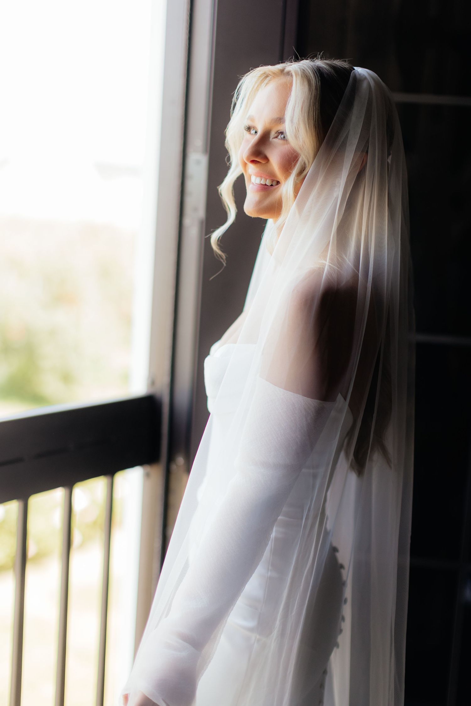 Bride in white gown stands near window with soft natural light filtering through.