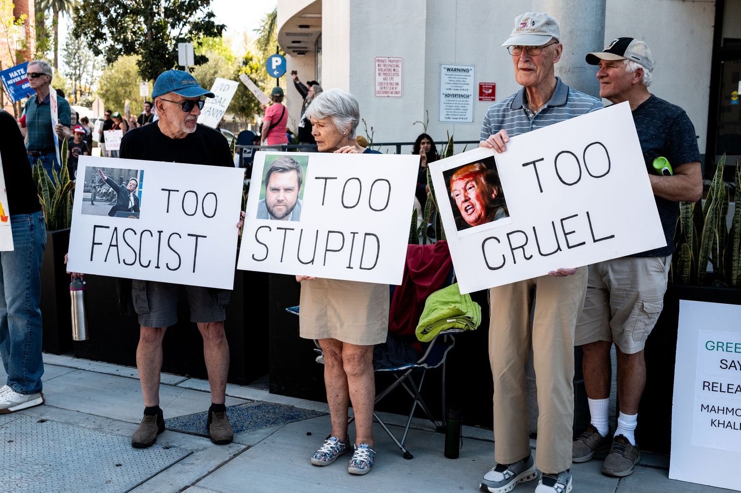 A line of protesters hold signs with critical messages at a public demonstration.