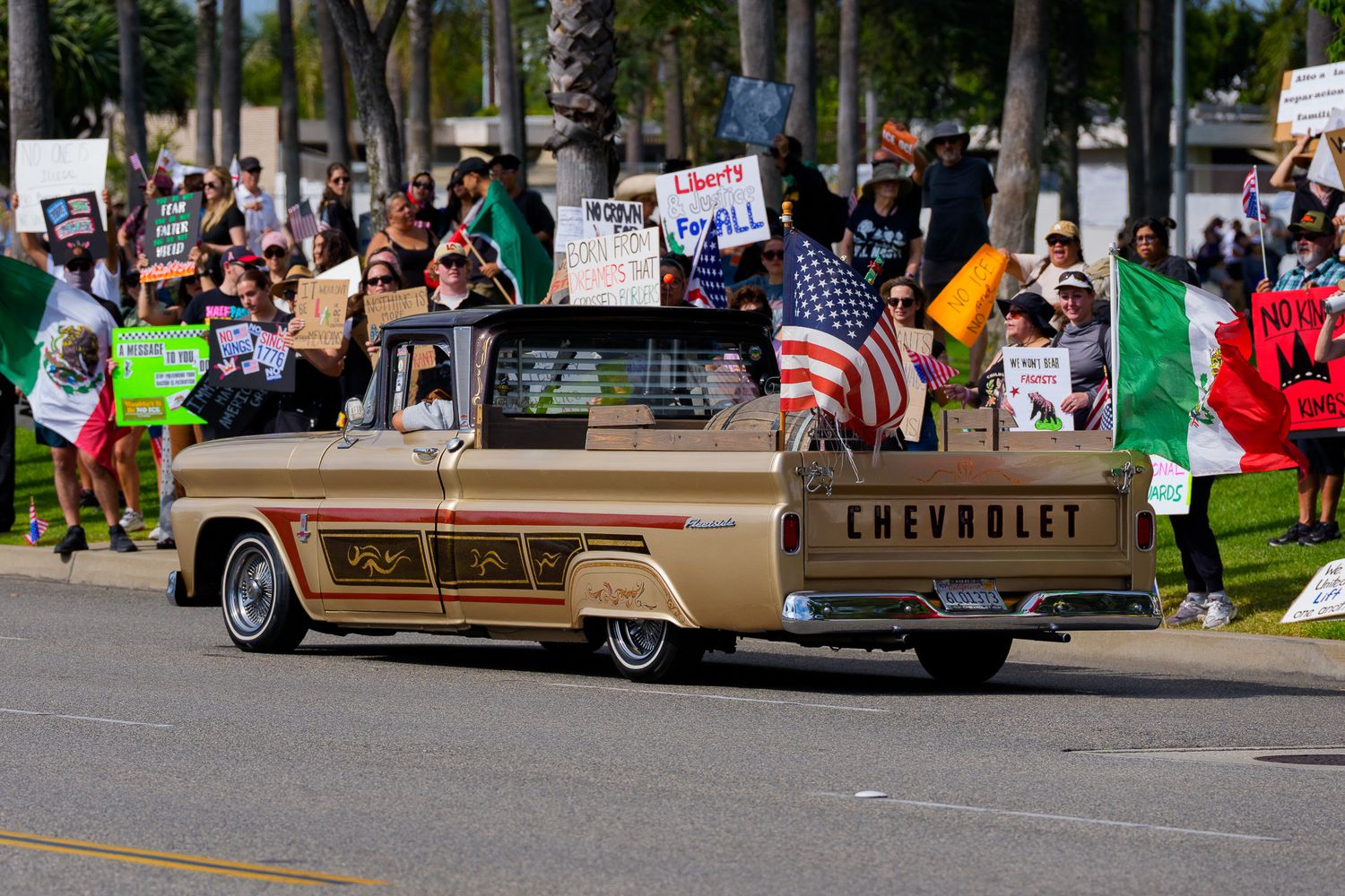 Vintage Chevrolet cars line up at an outdoor event with protesters and flags visible.