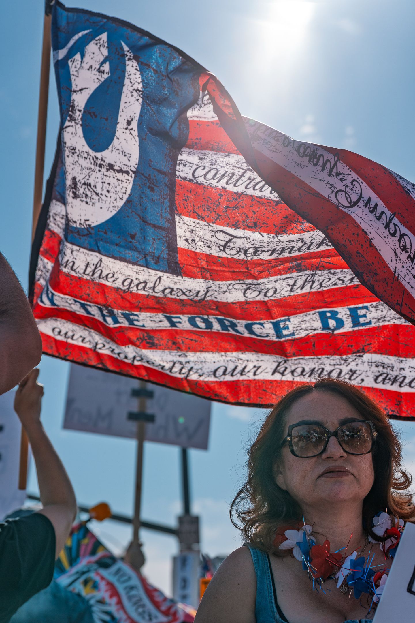 American flag with text waves against a bright blue sky during a protest.