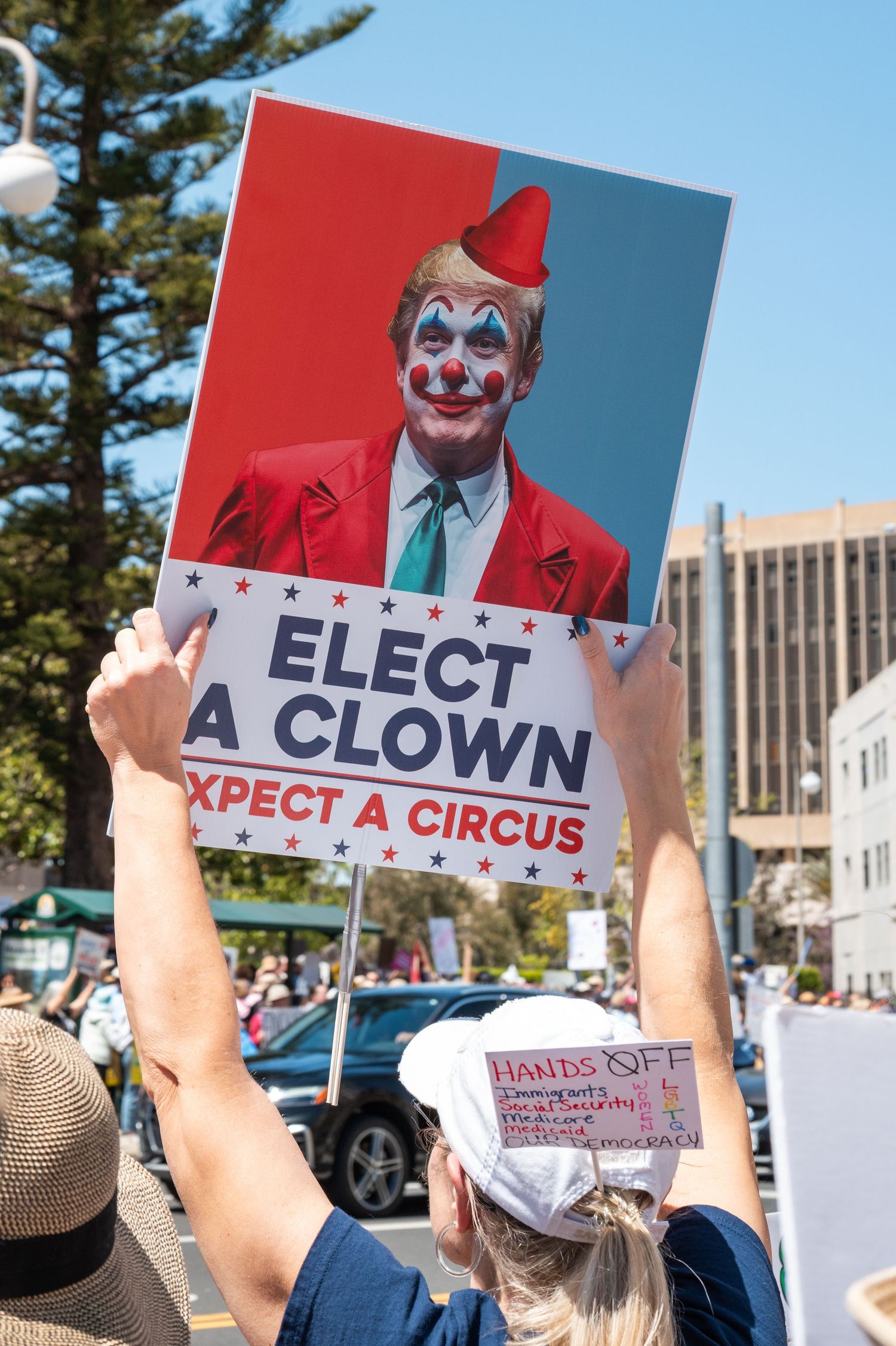 Protesters hold up signs featuring a clown-themed political message at an outdoor demonstration.