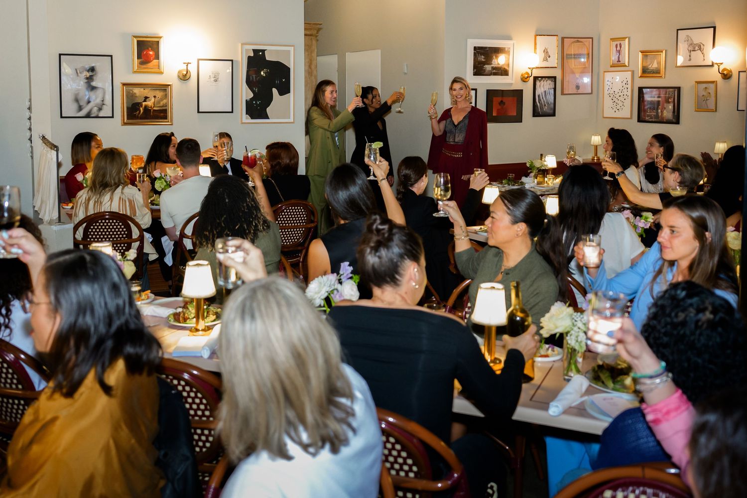 Crowded restaurant event with women dining at tables, raising glasses, and socializing in a warmly lit venue.
