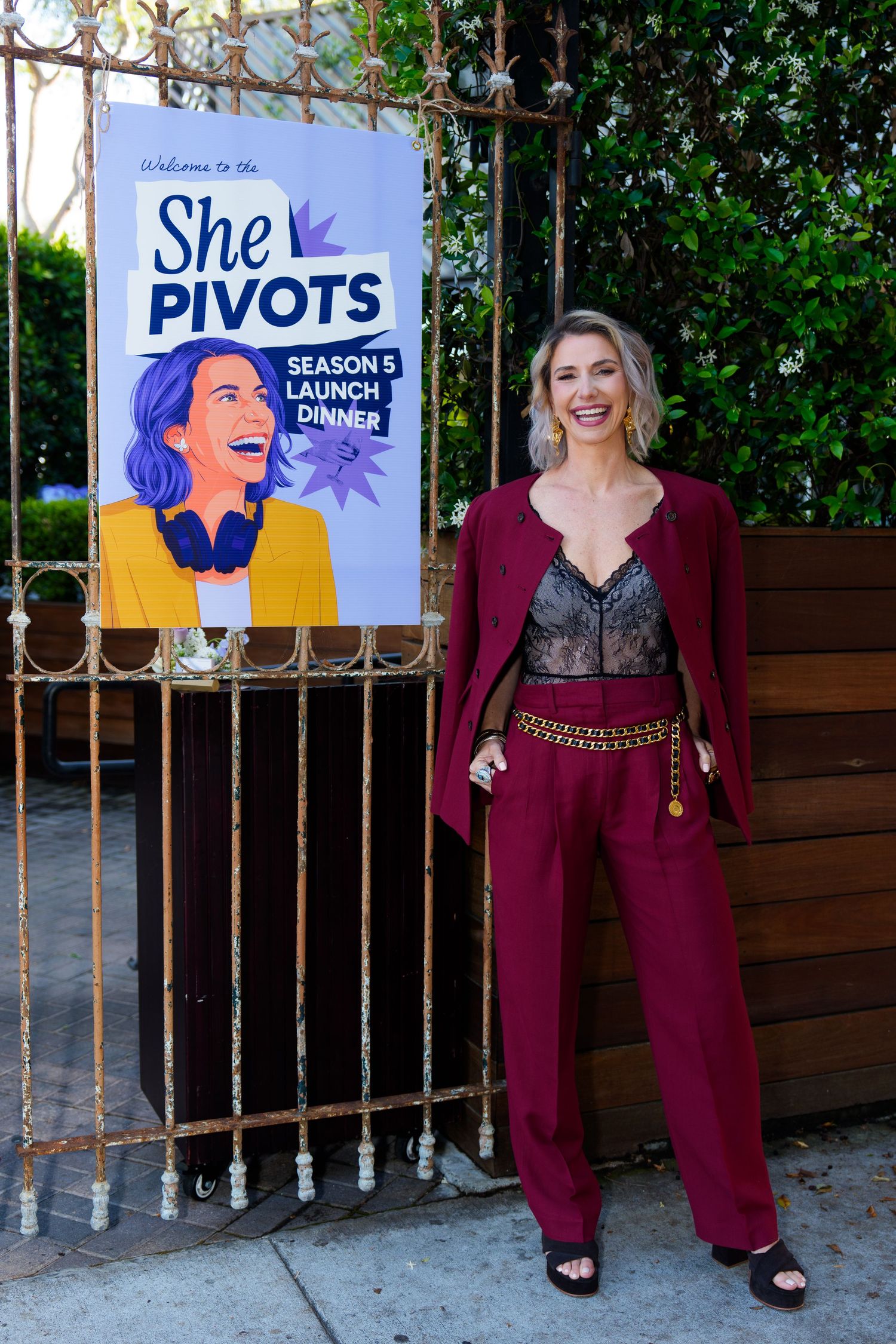 Woman in burgundy suit poses beside She Pivots Season 5 Launch Dinner sign outdoors.