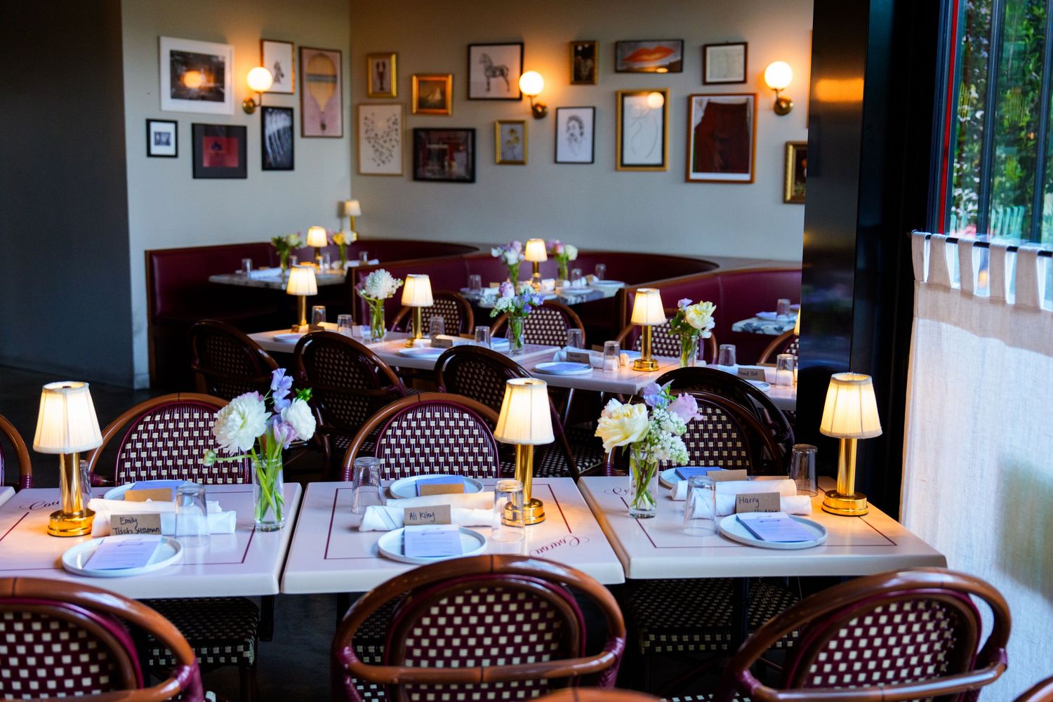 Elegant restaurant dining room with wicker chairs, wooden tables, glowing lamps, floral centerpieces, and art-covered walls.