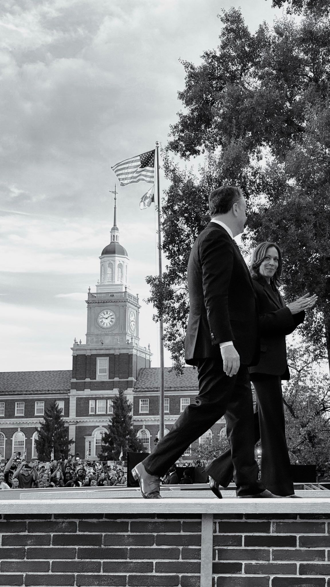 Black and white photo of people walking on a brick wall with a historic clock tower and American flag in the background.